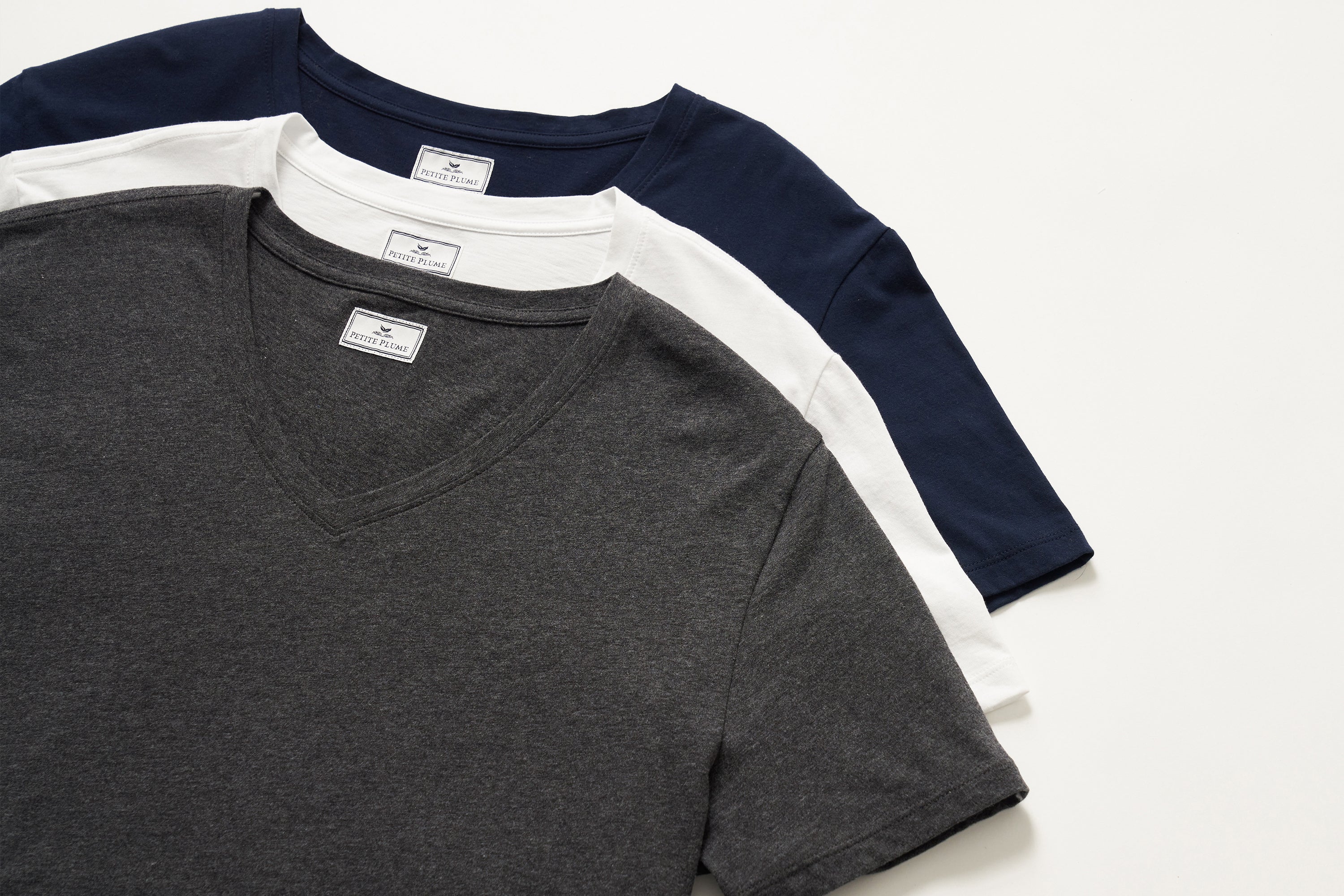 Three neatly stacked t-shirts in dark gray, white, and navy blue, each with a visible collar and neck tag, displayed on a plain white background.
