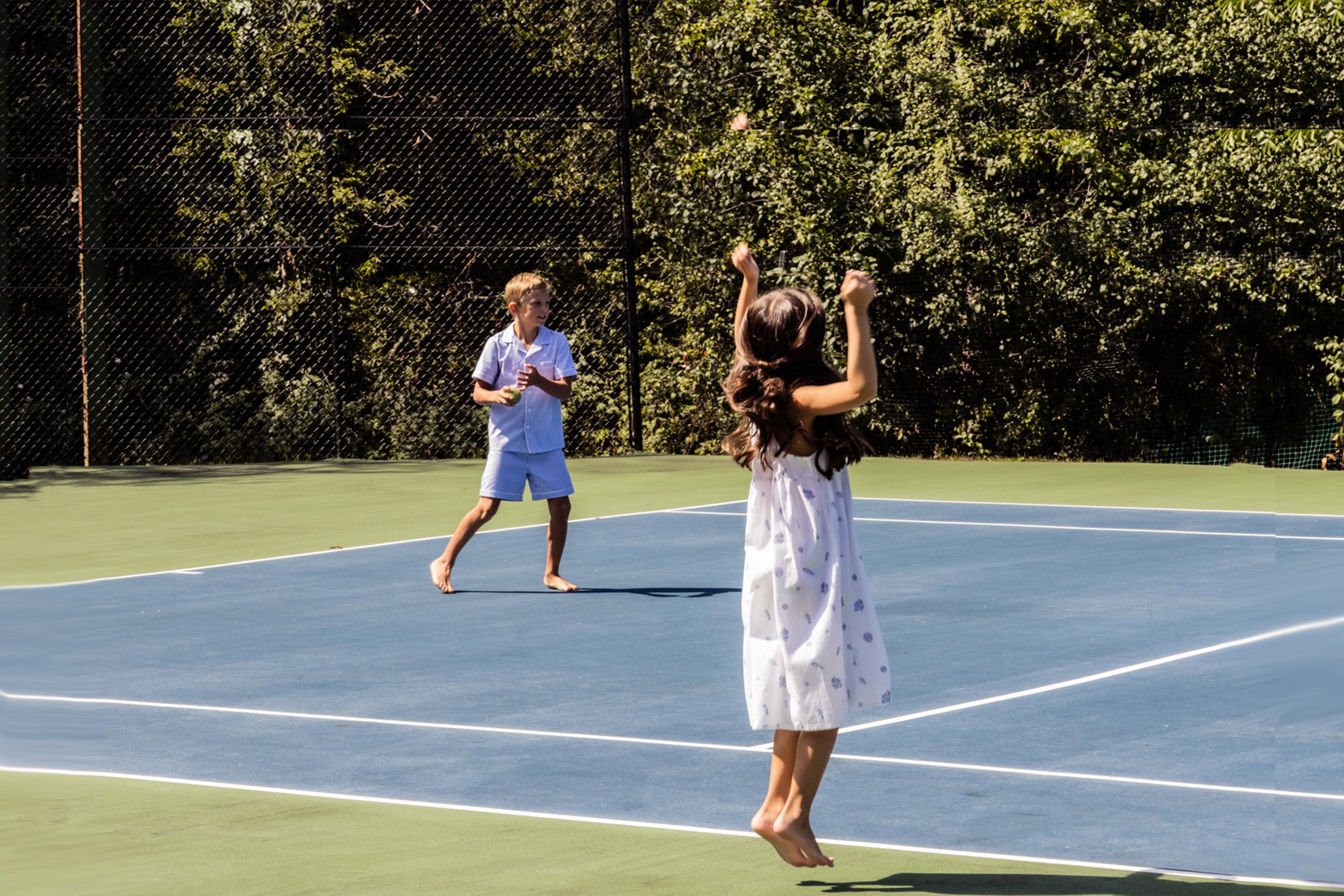 Two children play barefoot tennis on an outdoor court. The girl in a white dress jumps to hit the ball while the boy in a blue outfit watches, surrounded by trees and sunshine.
