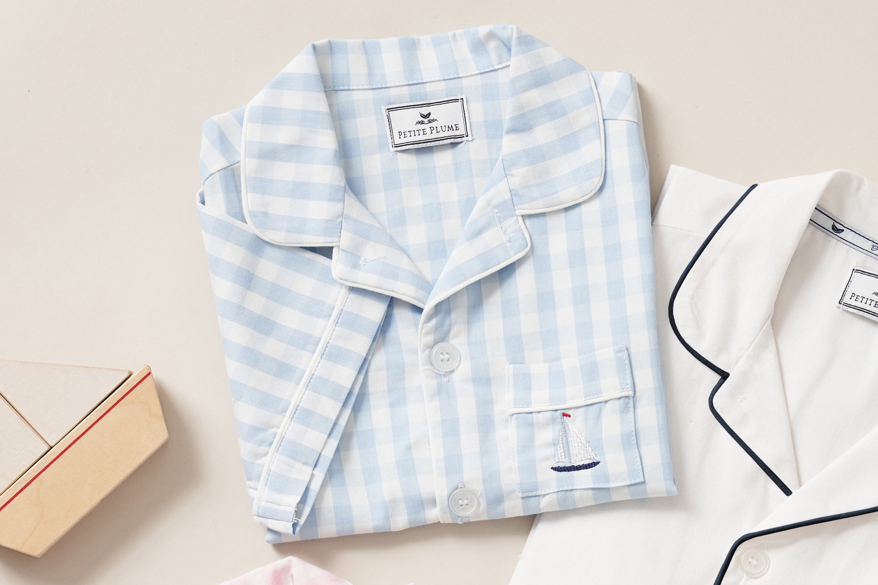 Folded childrens pajamas: one set in light blue gingham with a small sailboat embroidered on the pocket, and another in white with dark piping, both displayed on a neutral background.