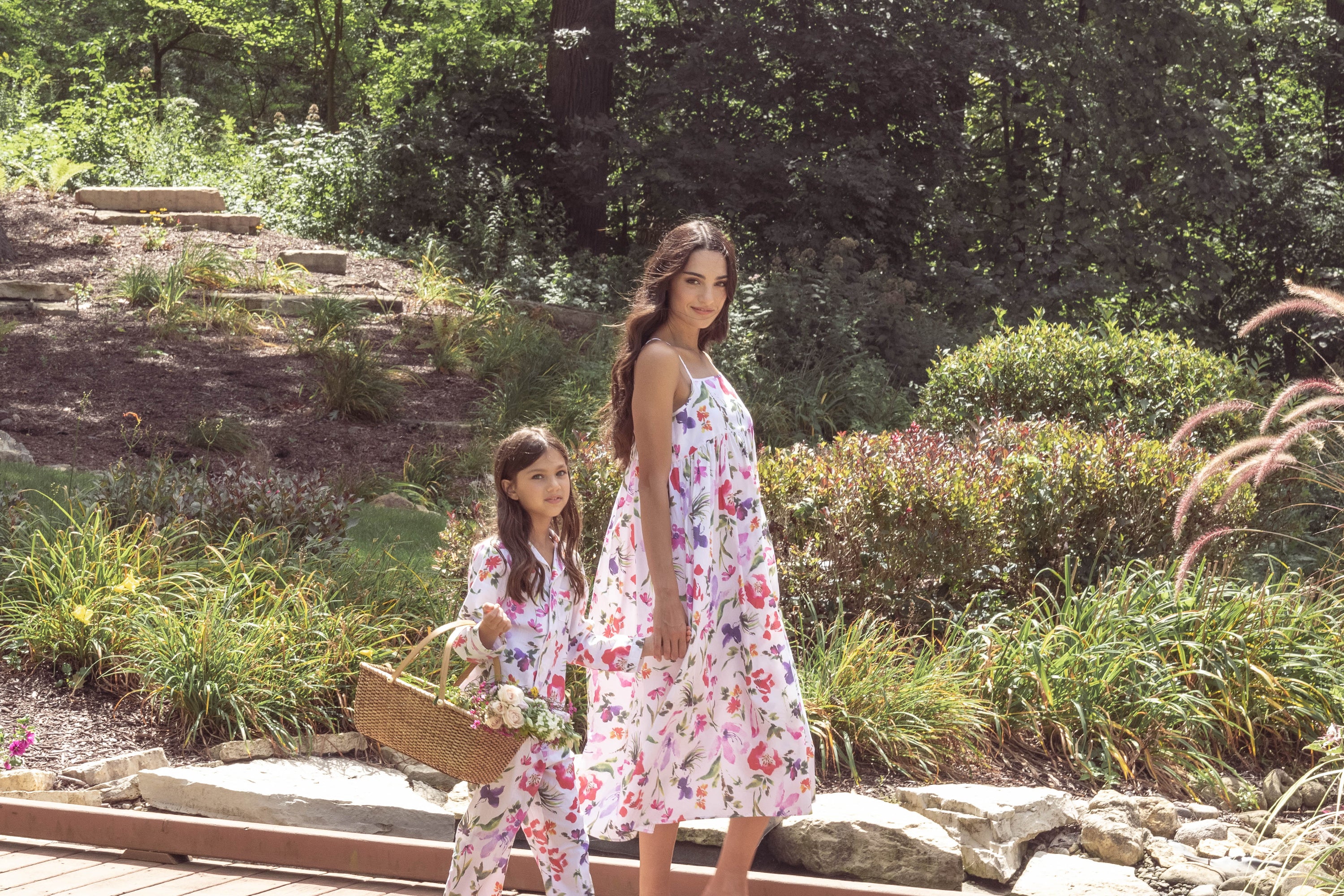 A woman in a floral dress and a young girl in matching pajamas walk outdoors; the girl carries a basket of flowers. They are surrounded by greenery and stone landscaping.
