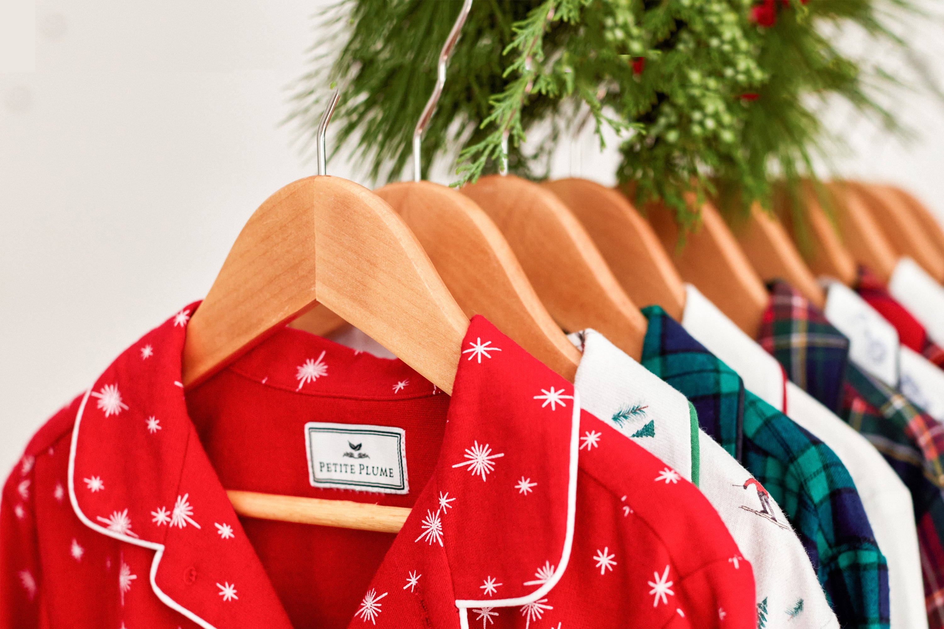 A row of colorful pajamas on wooden hangers, including red, white, and plaid patterns, hanging beneath green holiday foliage.
