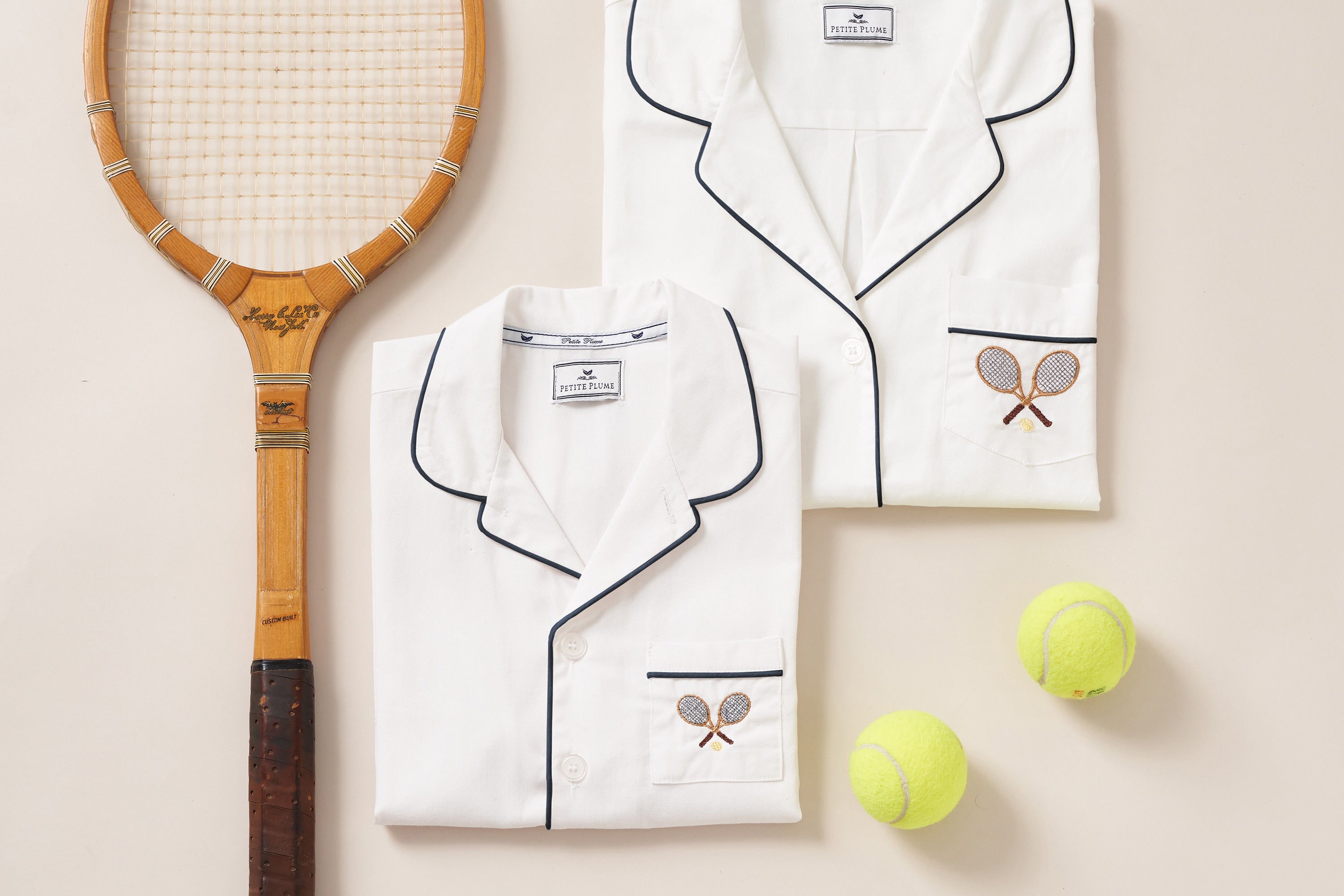 Two folded white pajama tops with black piping and embroidered tennis rackets, next to a vintage wooden tennis racket and two yellow tennis balls on a cream background.