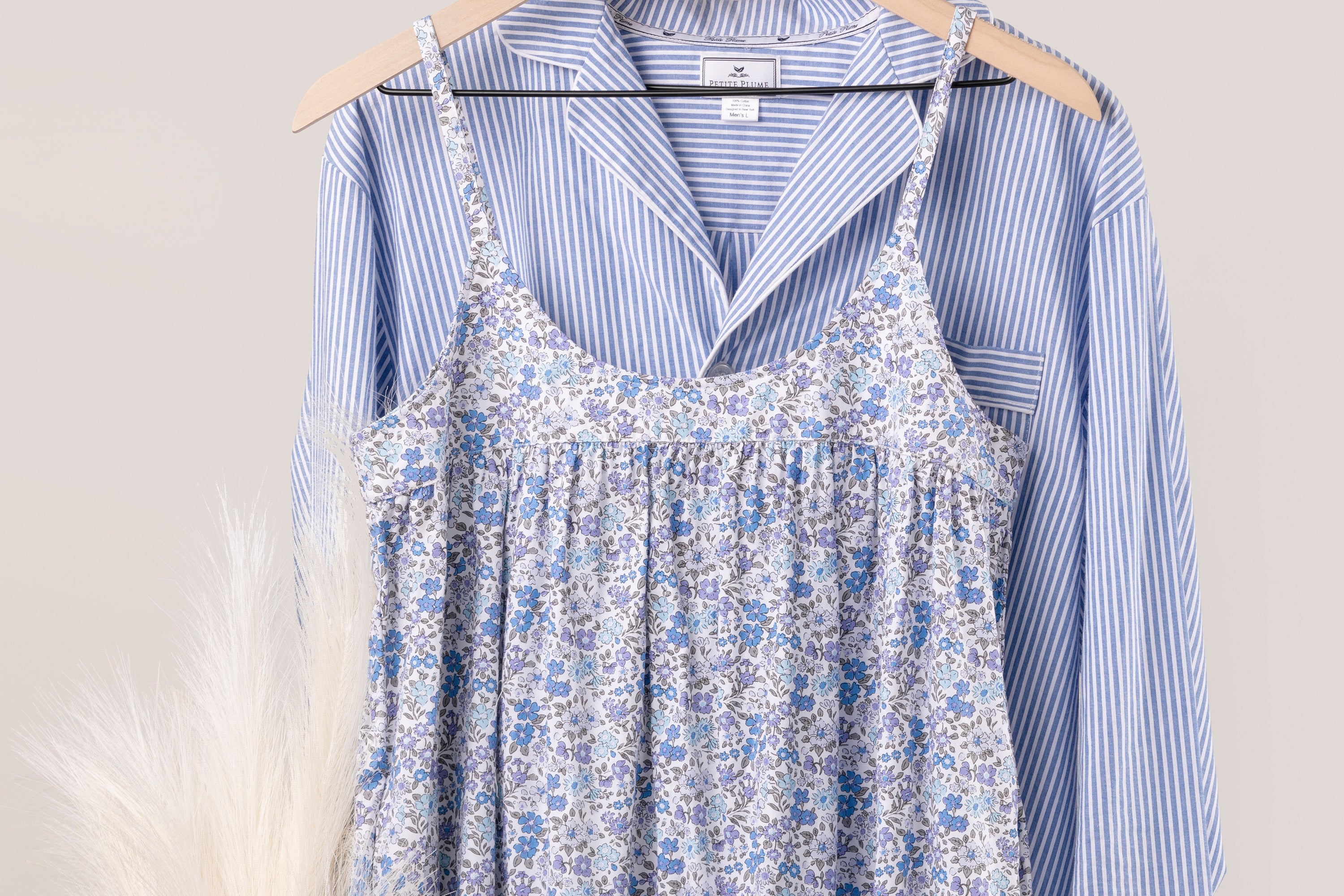 A blue and white striped button-up shirt hangs on a hanger, layered underneath a sleeveless, floral-patterned dress in shades of blue, white, and purple. Pale decorative grass is visible to the left.