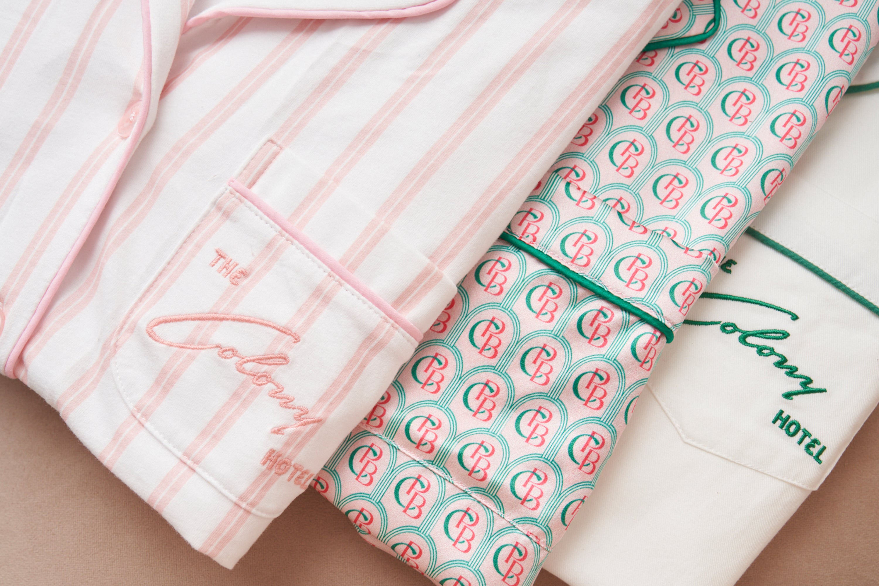 Three folded pajama tops: one pink striped with “The Colony Hotel” embroidered, one with a pink and green circular pattern, and one white with green trim and “The Colony Hotel” embroidery.