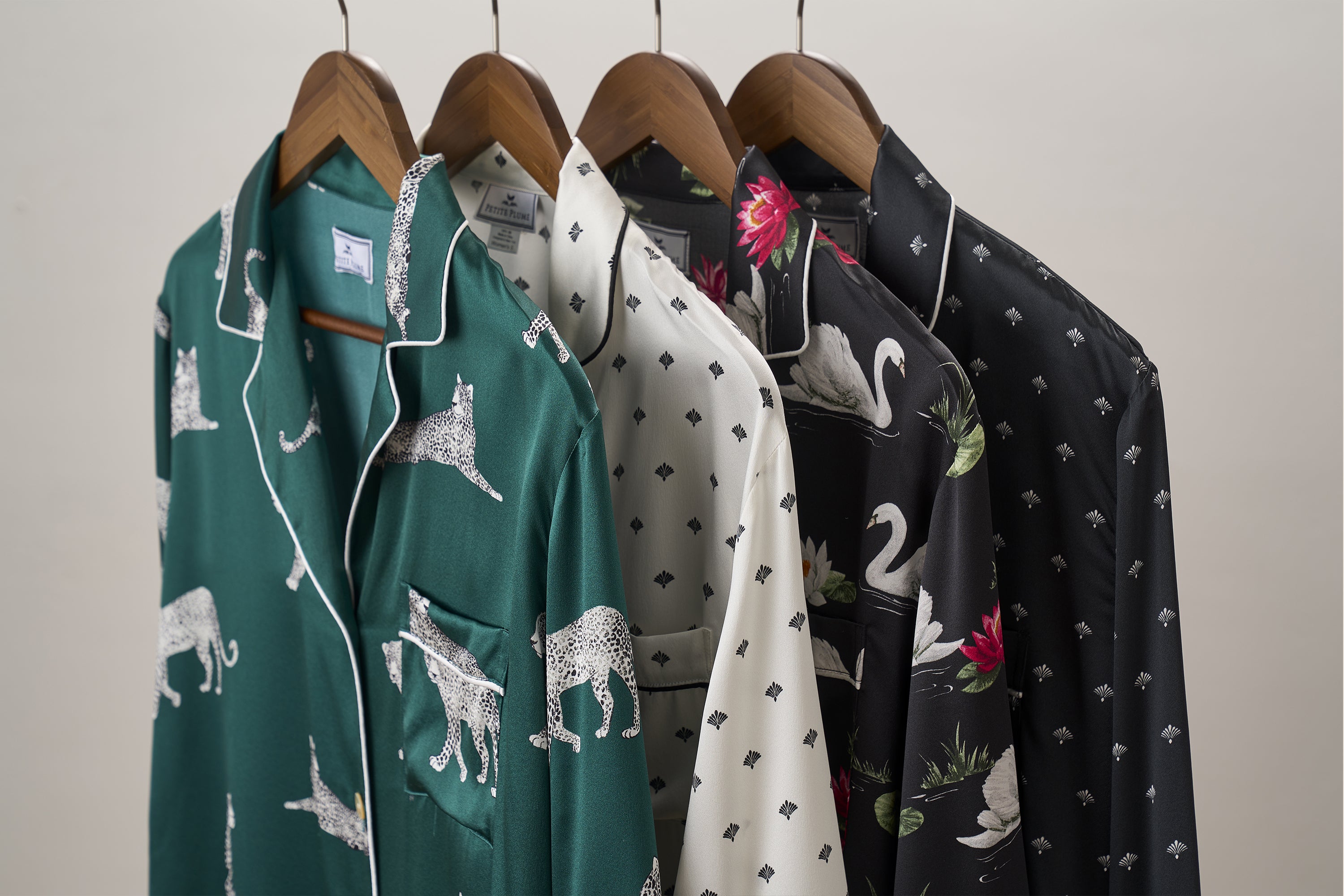 Four patterned pajama shirts on wooden hangers: green with cheetahs, white with small black diamonds, black with swans and red flowers, and black with small white diamonds, all against a plain background.