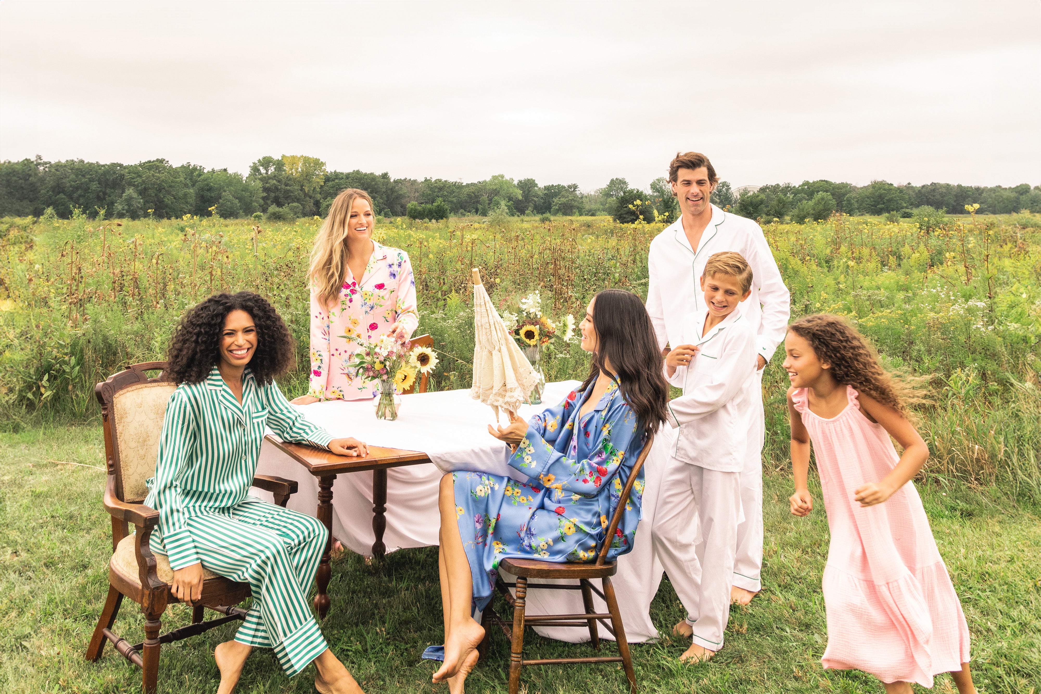 Five people in colorful pajamas gather around a table with sunflowers and a sailboat centerpiece, outdoors in a grassy field under a cloudy sky, smiling and enjoying each others company.