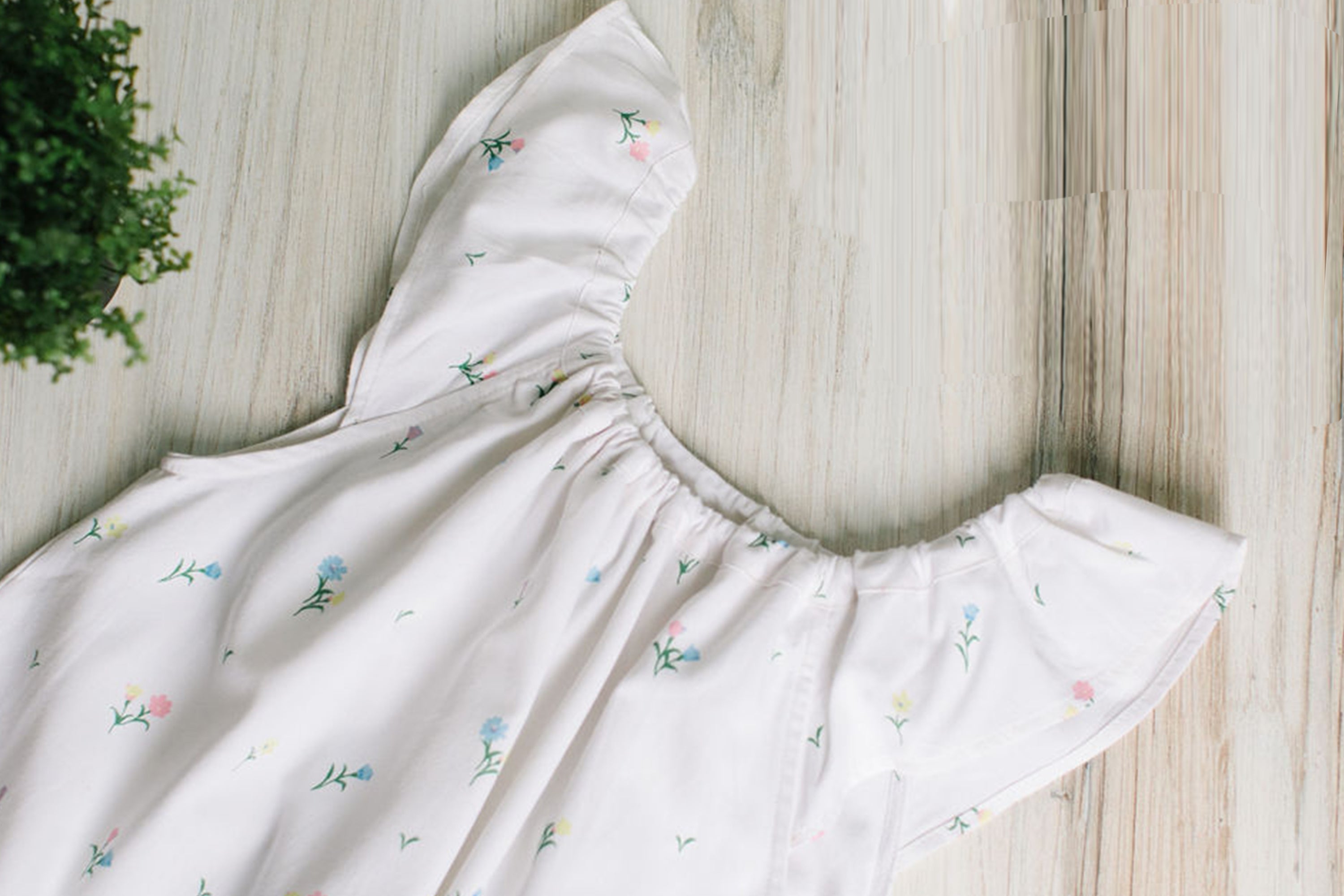A white dress with small, colorful floral prints and ruffled sleeves is laid out on a light wooden surface next to a green potted plant.