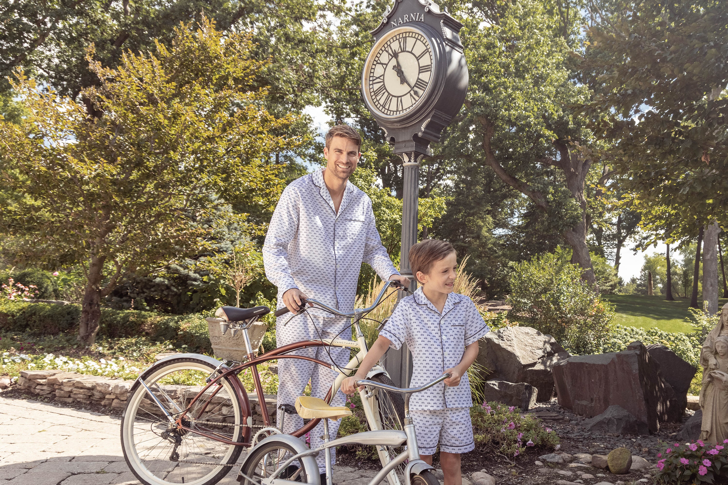 A man and a boy wearing matching pajamas stand by vintage bicycles near a large outdoor clock labeled Narnia, surrounded by greenery and trees on a sunny day.
