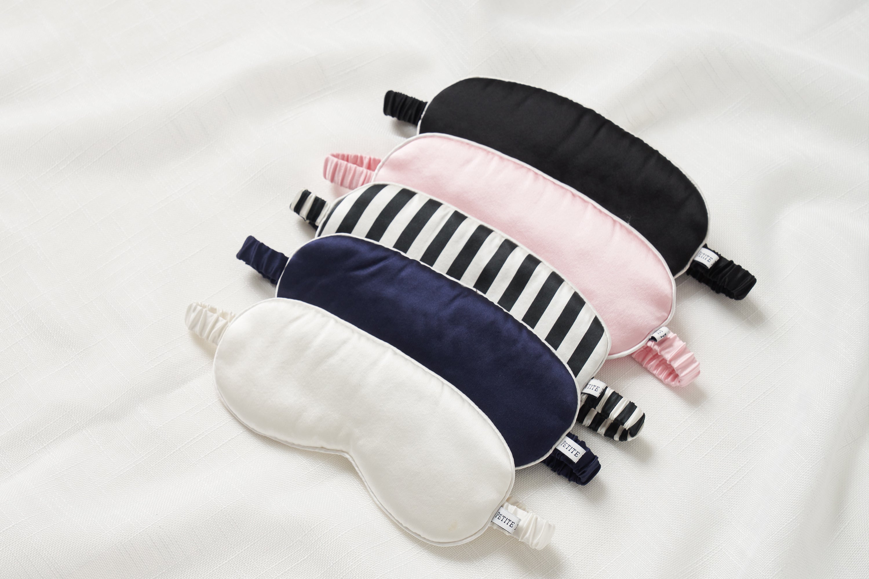 Five sleep masks in different colors and patterns—black, pink, black and white stripes, navy blue, and white—are arranged in a neat, overlapping row on a white fabric background.