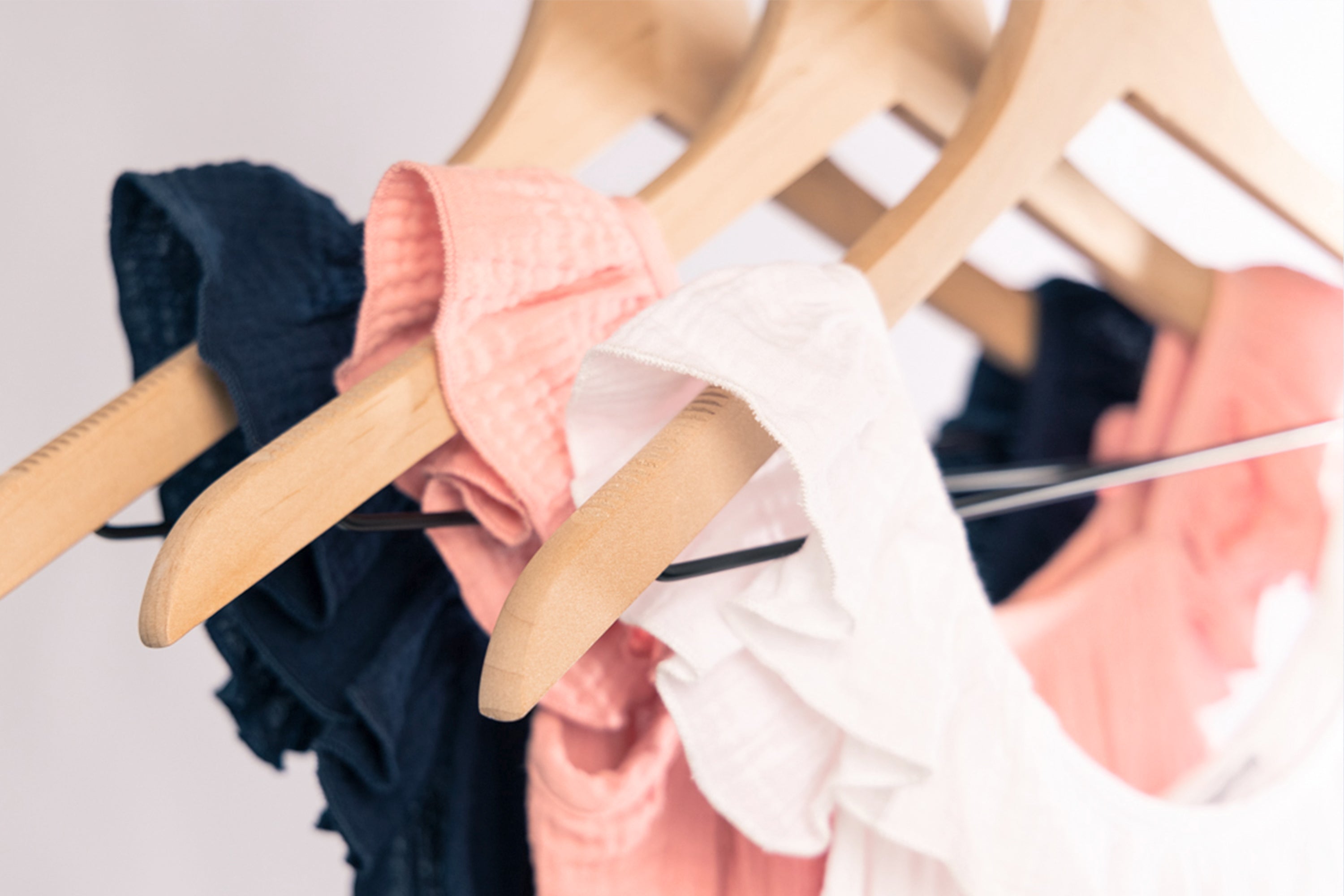Close-up of wooden hangers holding ruffled sleeveless tops in navy blue, pink, and white, hanging on a clothing rack against a soft, light background.
