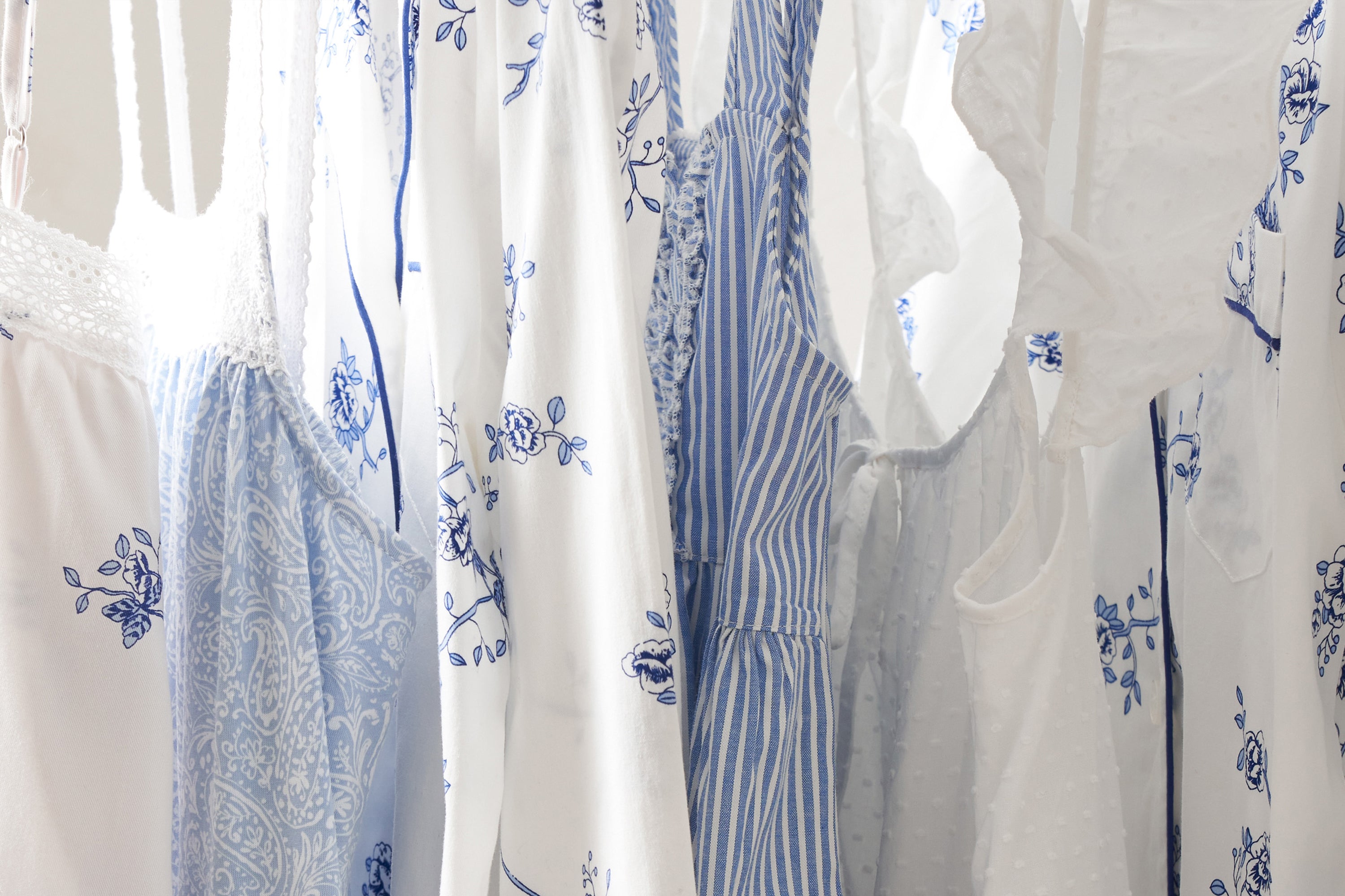 A collection of white and blue garments on hangers, featuring floral and stripe patterns. The dresses and blouses vary in texture and design, creating a summery, fresh appearance.