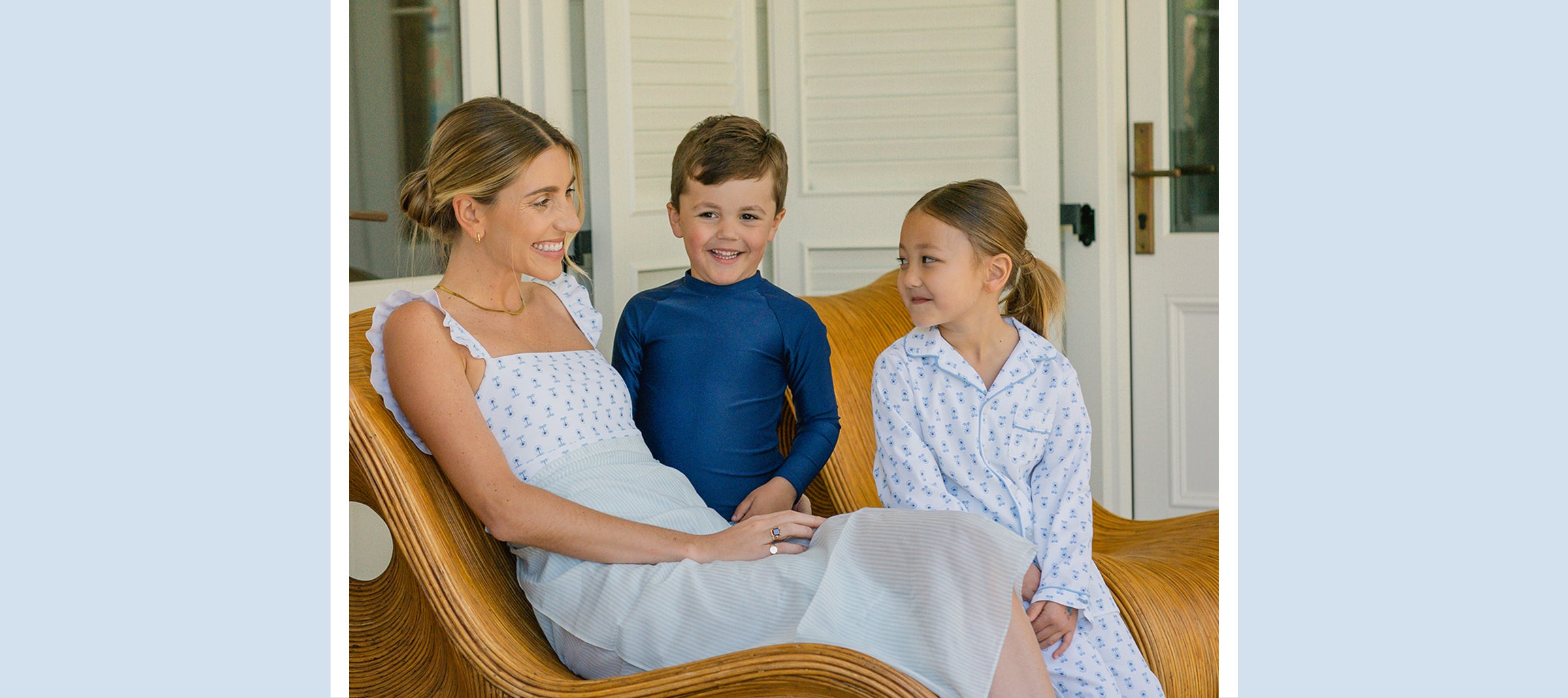 A woman sits on a wicker chair smiling at two young children, a boy in a navy rash guard and a girl in a white patterned dress, all enjoying time together on a porch.