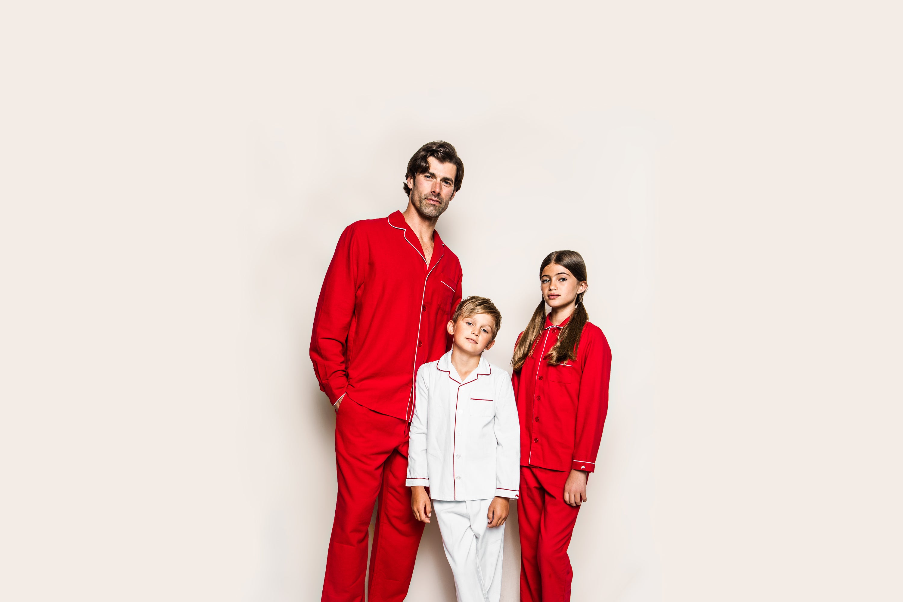 A man and a girl in matching red pajamas stand beside a boy in white pajamas with red trim, all posing against a plain light background.