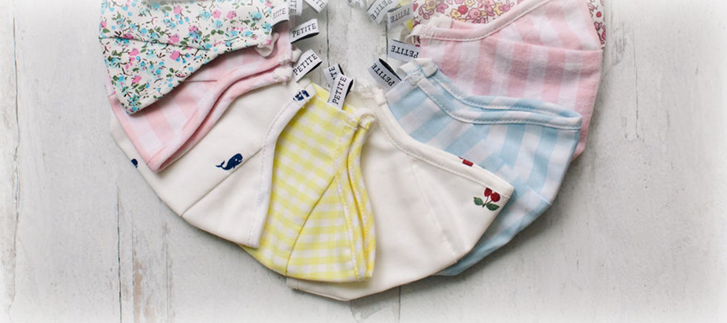 A semicircle arrangement of pastel-colored and patterned cloth face masks with small label tags, displayed on a white wooden surface.