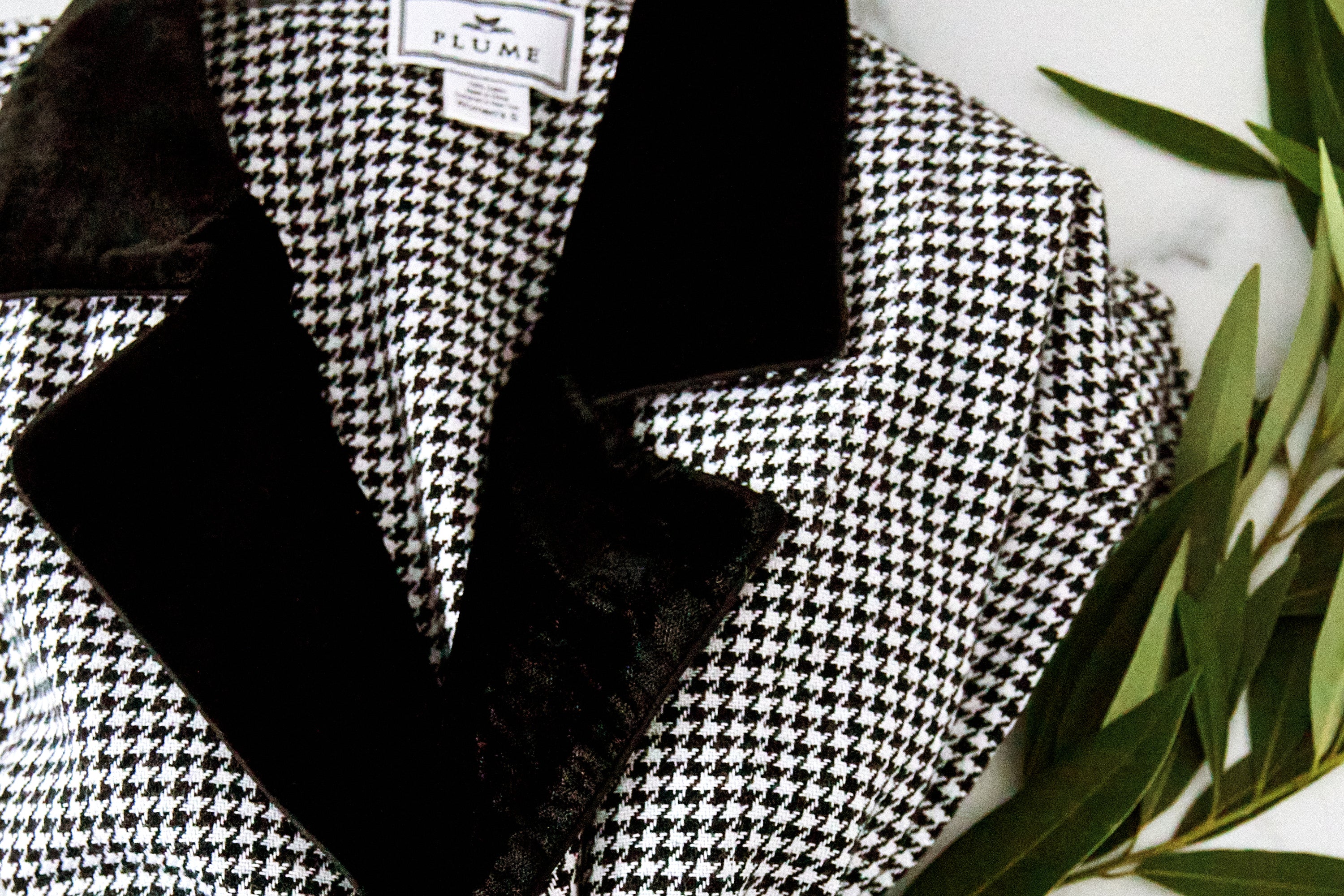 Close-up of a black and white houndstooth patterned jacket with a black velvet collar, lying next to green leafy branches on a white surface.