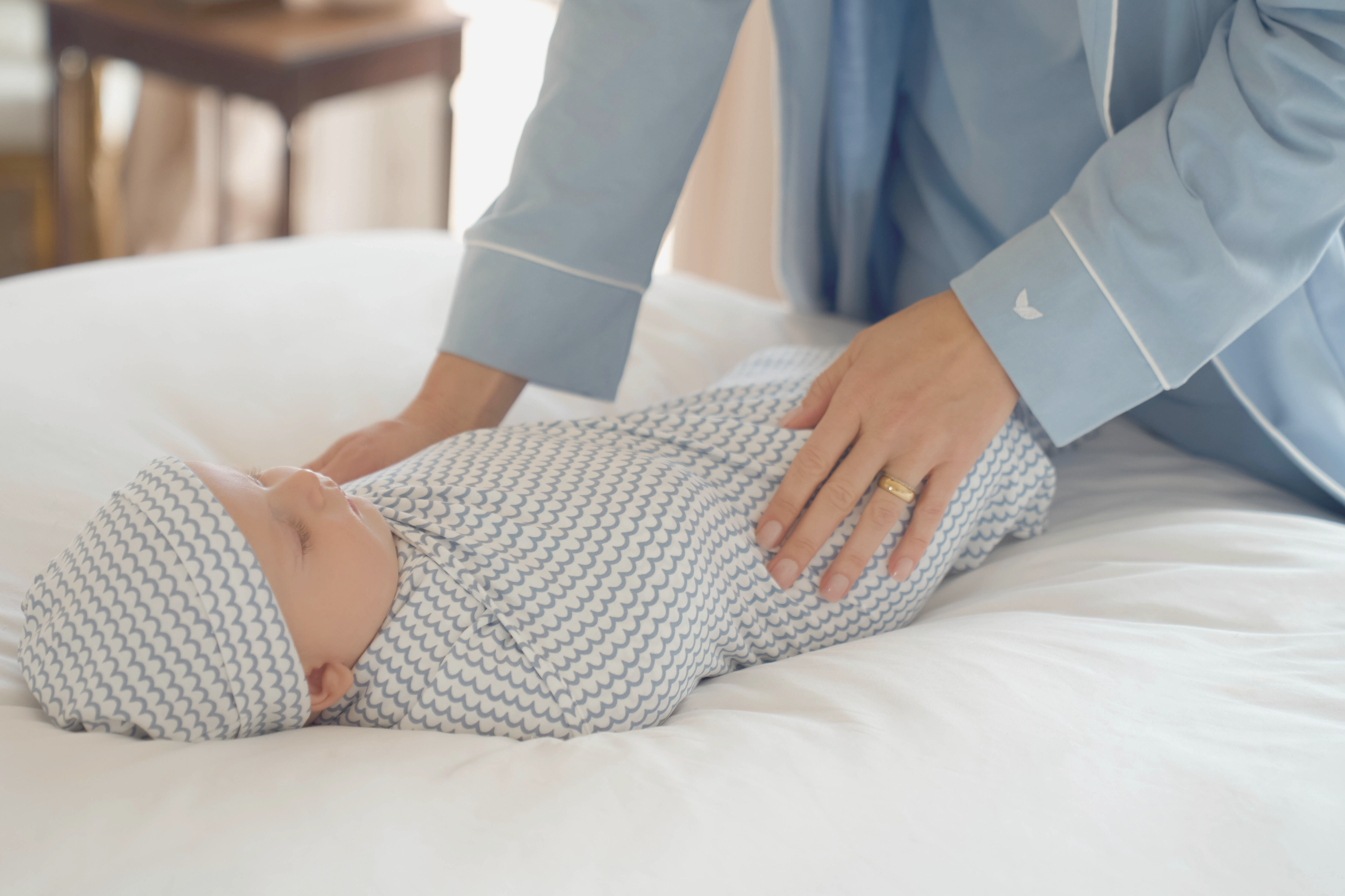 A person in blue pajamas gently places their hands on a swaddled baby lying on a white bed. The baby is wearing a matching hat and patterned swaddle blanket.
