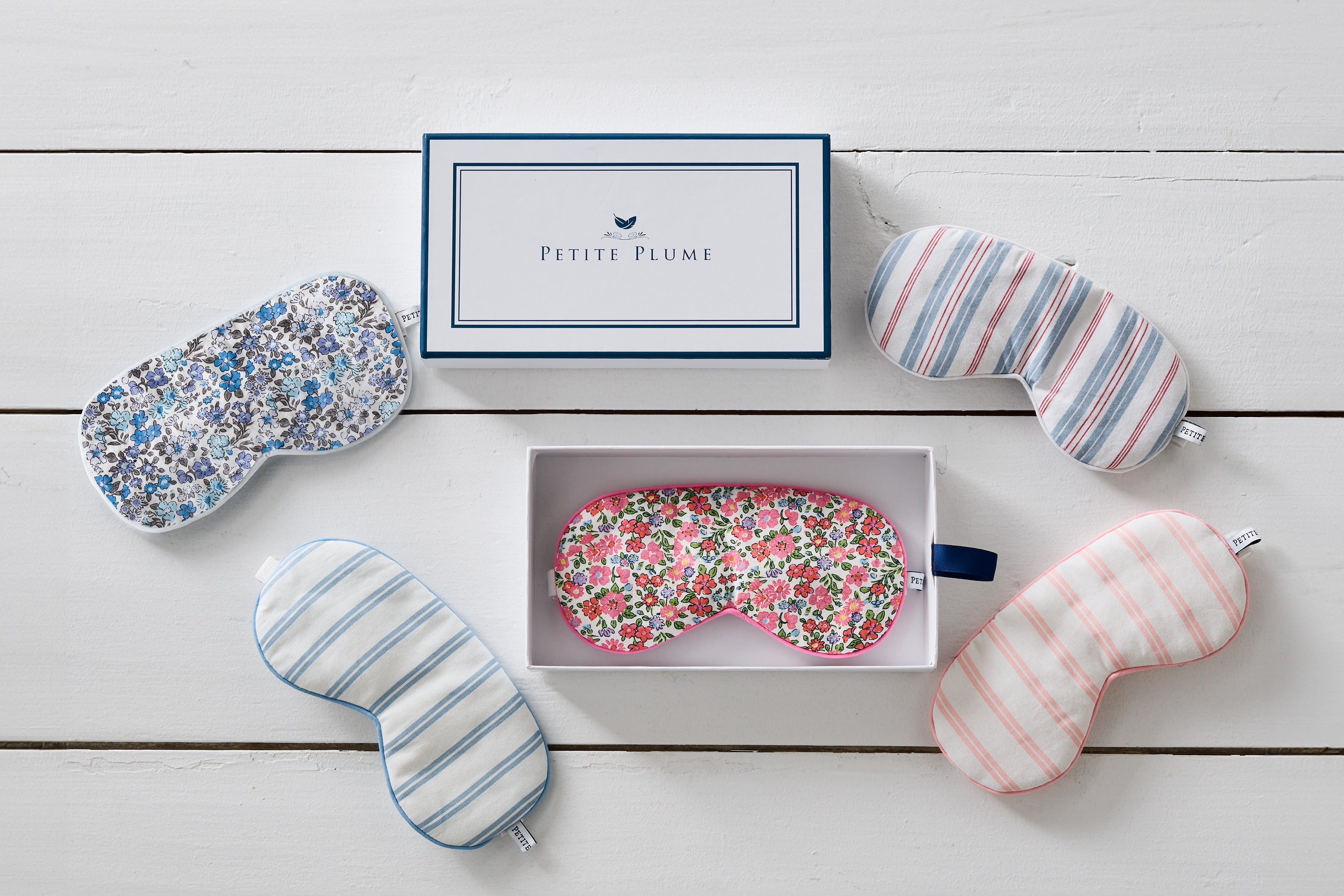 Five patterned sleep masks are arranged around an open Petite Plume box on a wooden surface. The box contains one sleep mask with a floral design matching the others, including blue, stripe, and floral patterns.