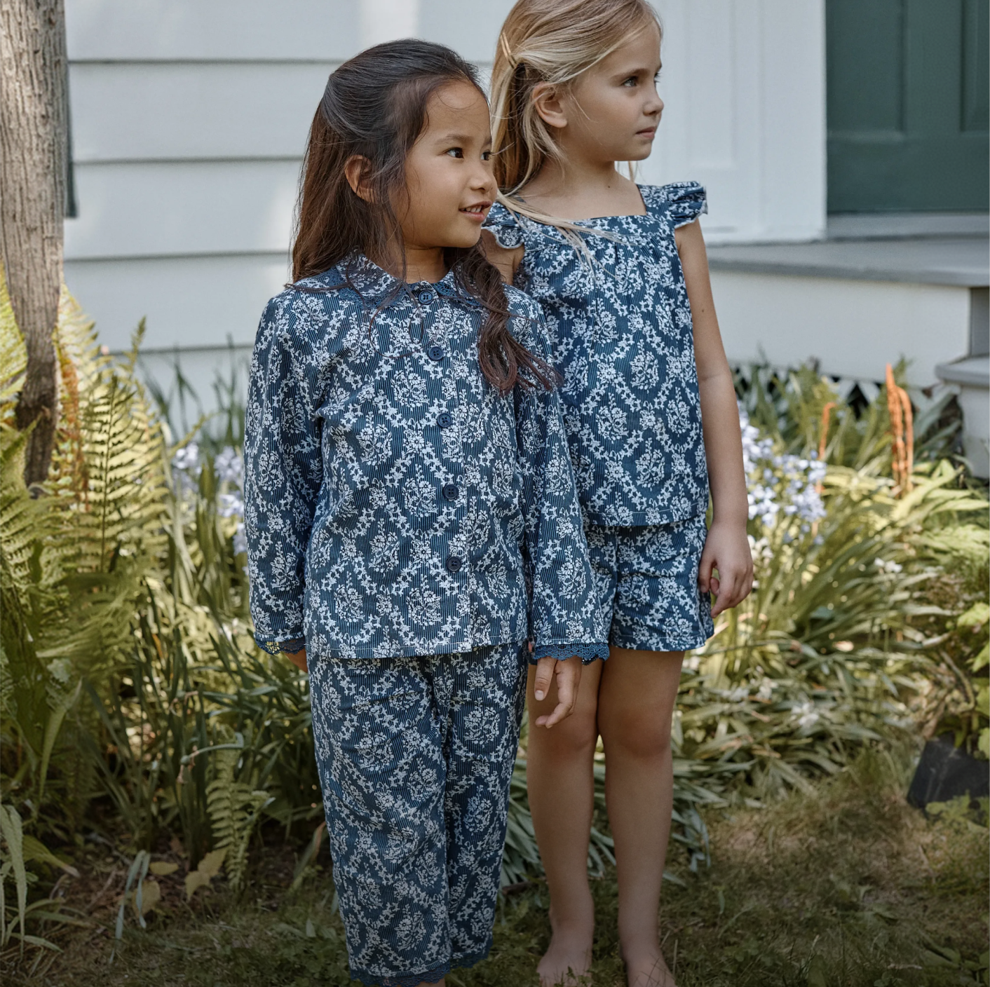 Two young girls stand outside in a garden, both wearing matching blue and white floral patterned outfits—one in long sleeves and pants, the other in a short-sleeved top and shorts. Ferns and flowers grow nearby.