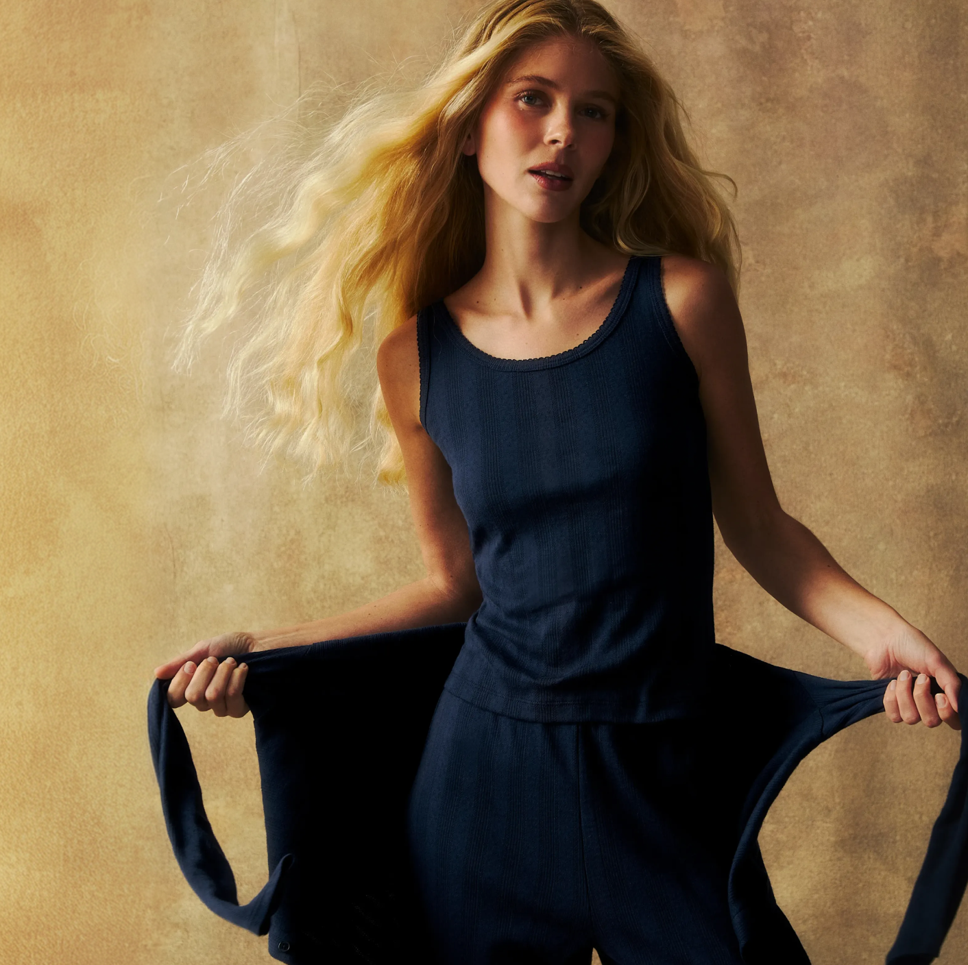 A woman with long blonde hair wears a navy tank top and pants, holding out a matching jacket. She stands against a warm, textured beige background, looking slightly past the camera.