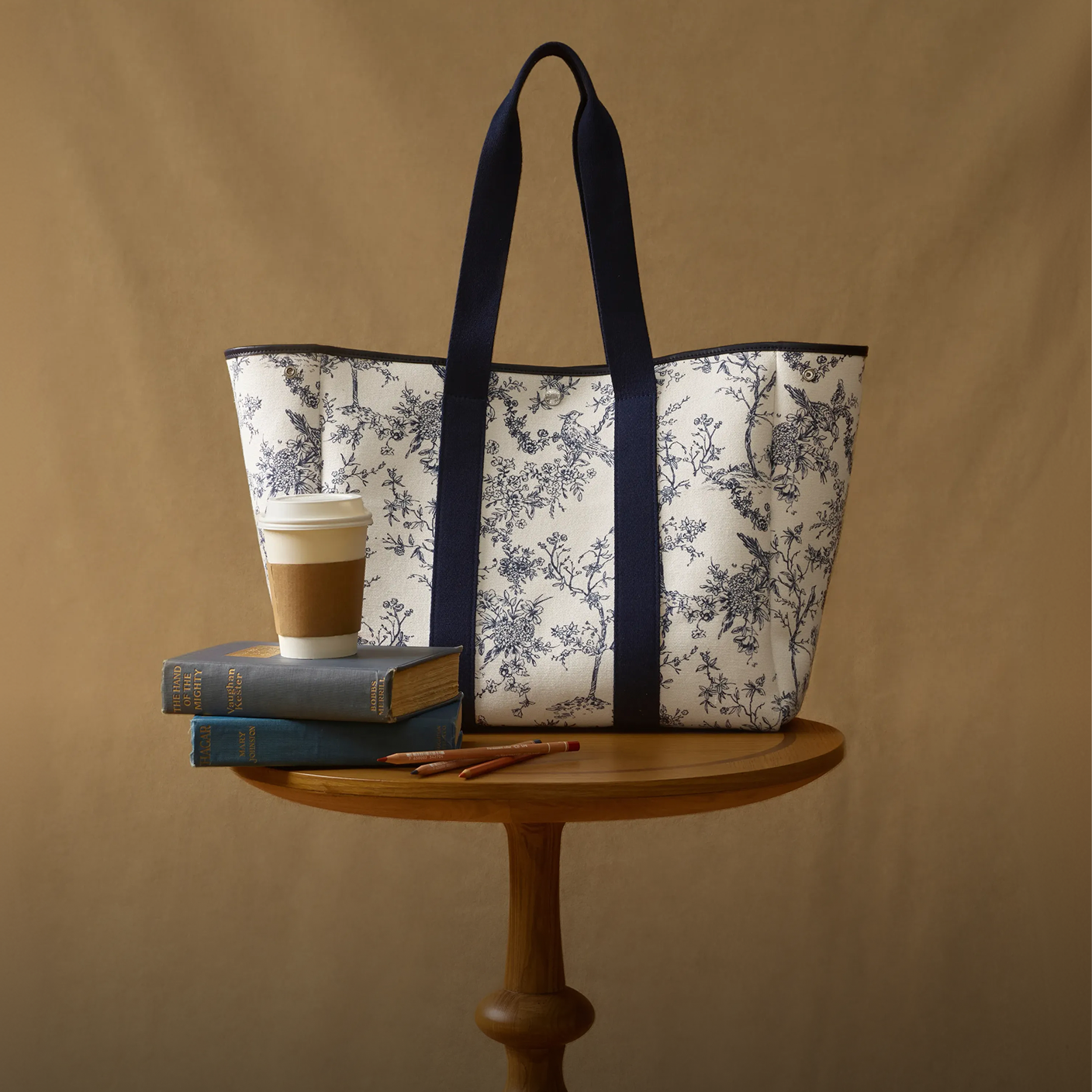 A floral-patterned tote bag with dark handles sits on a round wooden table with two books, a coffee cup, and two pencils in front of a plain brown background.