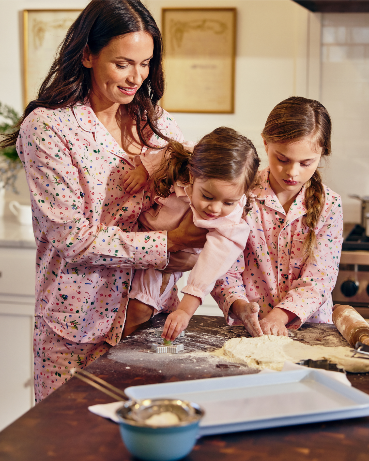 A woman and two young girls, all in matching pink pajamas, bake together in a kitchen. One girl shapes dough with cookie cutters while the woman holds the younger child, who also reaches for the dough.