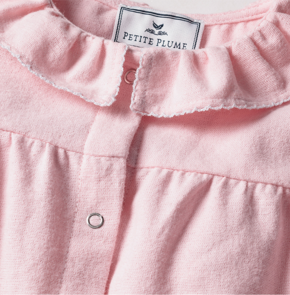 Close-up of a light pink ruffled collared garment with silver snap buttons and a label reading Petite Plume sewn at the neck. The fabric appears soft and cozy.
