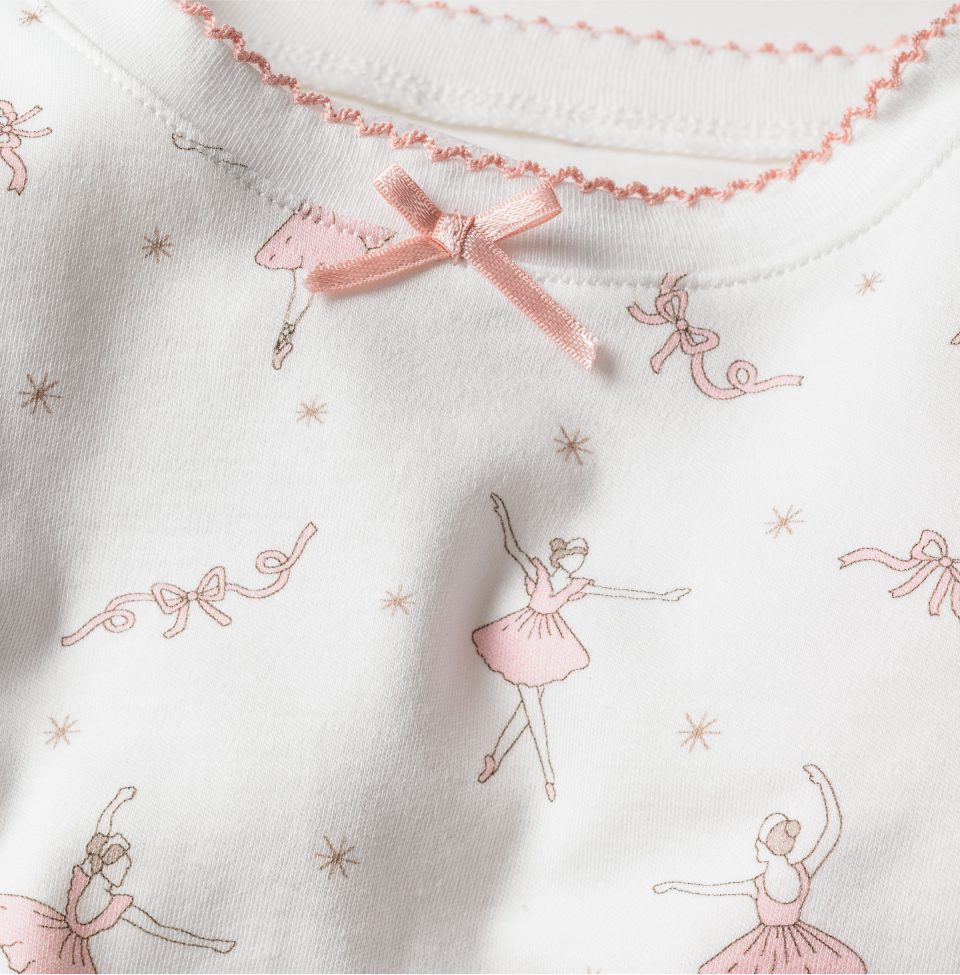 White fabric with light pink drawings of ballerinas and bows, featuring pink stitching along the neckline and a small pink satin bow at the collar center. The design has a delicate, whimsical, and feminine feel.