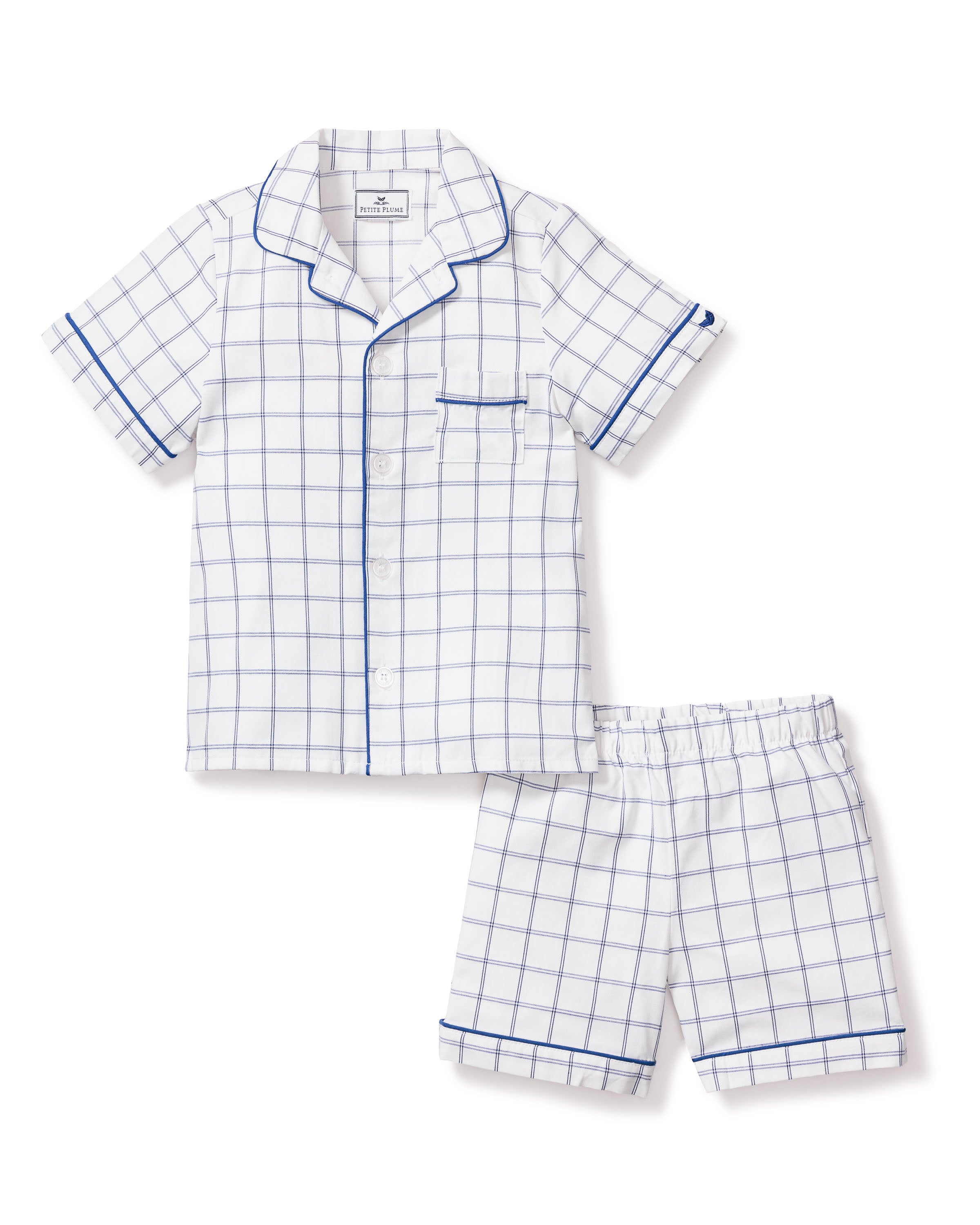 The Kids Twill Pajama Short Set in Nantucket Tattersall by Petite Plume features white short-sleeved pajamas with a blue grid pattern, including a button-up shirt with chest pocket and matching shorts. Blue piping accents add timeless detail to the collar, sleeves, pocket, and cuffs.