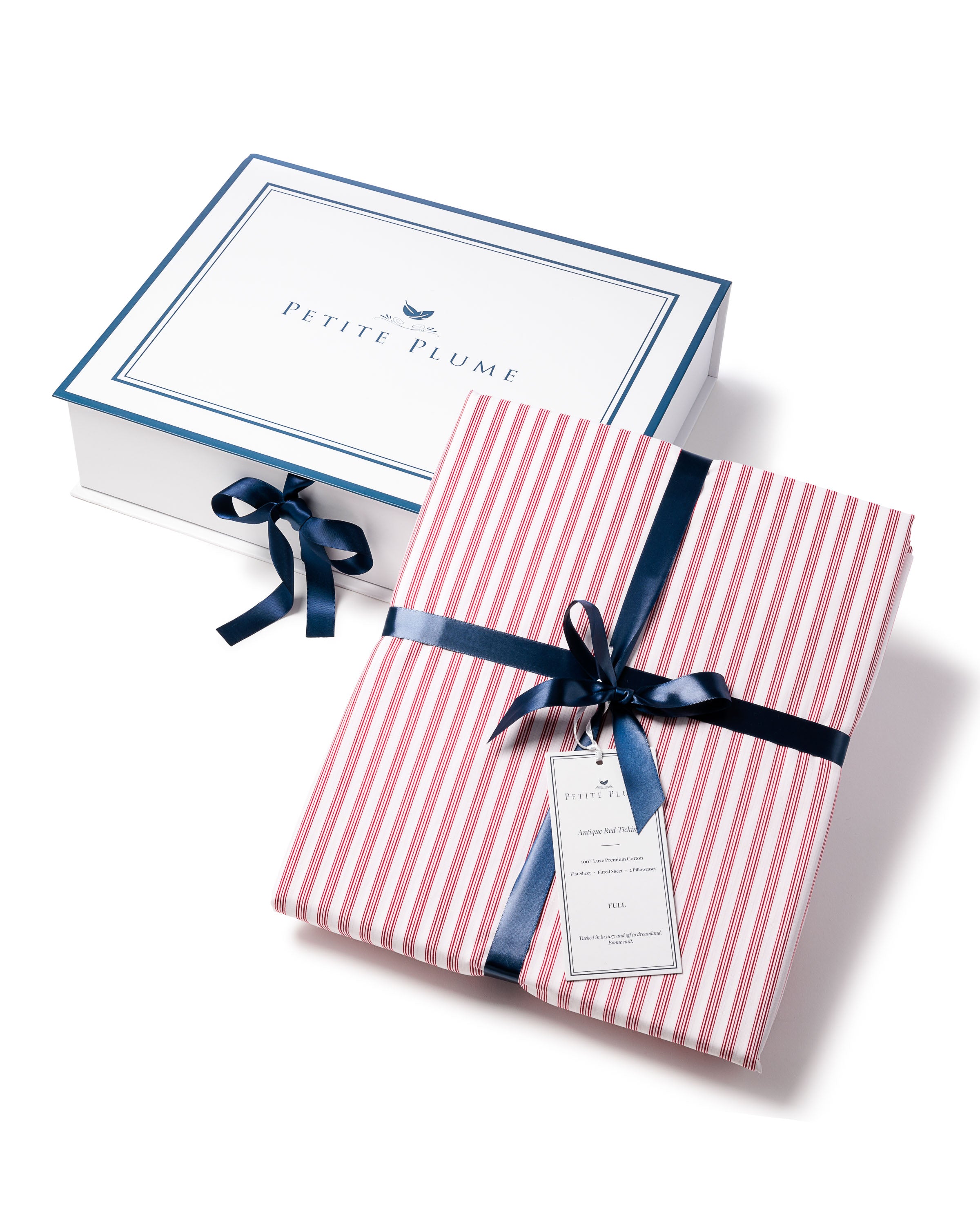 A folded Luxe Premium Cotton Bed Sheet in Antique Red Ticking by Petite Plume, resembling luxury bedding, is tied with a dark blue ribbon in front of an open white box. The box lid features a blue border and text. A tag dangles from the ribbon on this exquisite 100% cotton piece.
