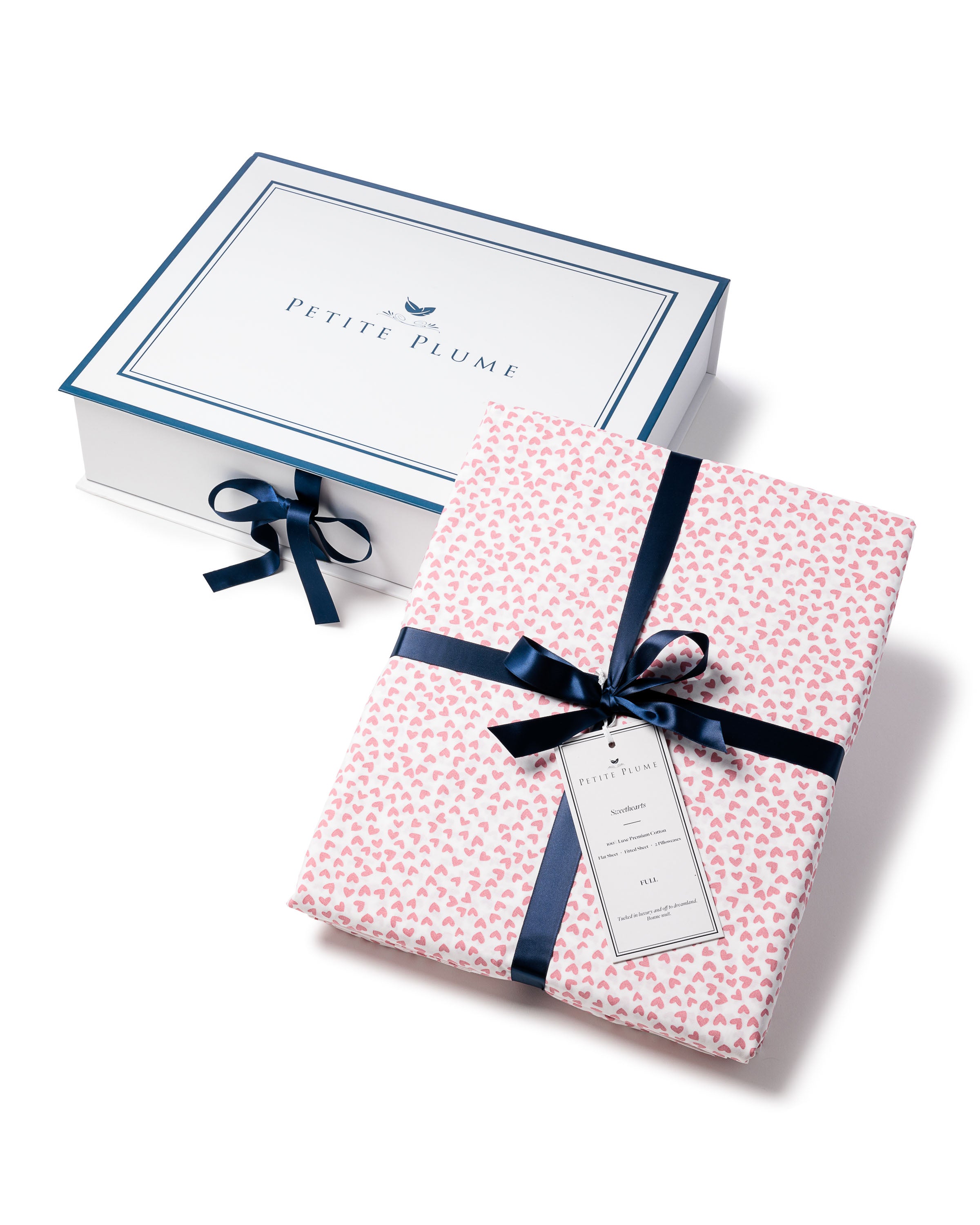 The image shows an open gift box with Petite Plume on the lid. Next to it is a set of Luxe Premium Cotton Sweethearts Bed Sheets, crafted from 100% cotton with pink heart-print fabric, neatly folded and tied with a dark blue ribbon and tag.