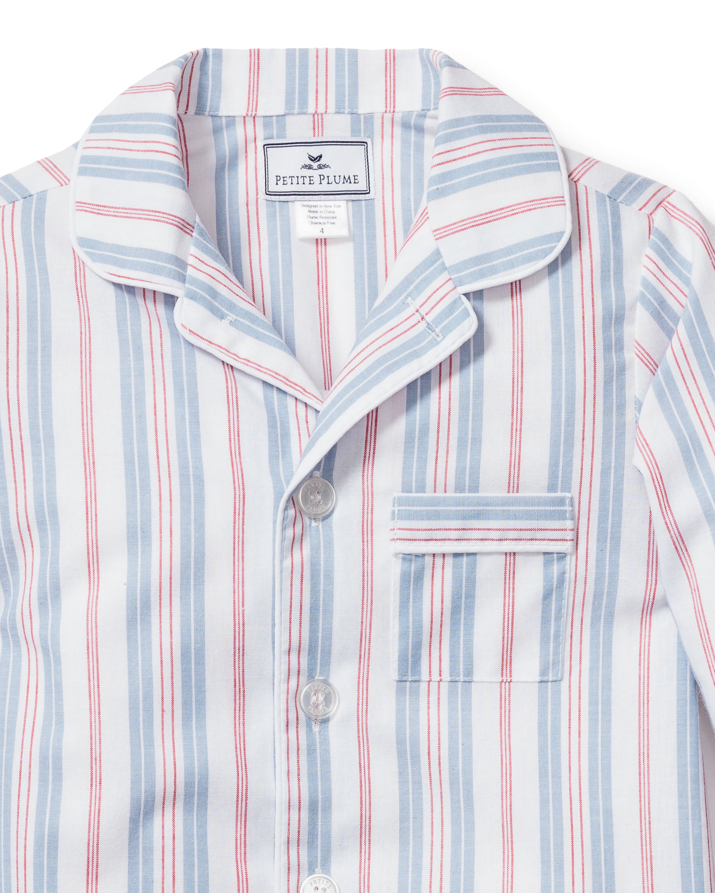 A close-up of the Kids Twill Pajama Short Set in Vintage French Stripes, featuring a collared top with blue and red vertical stripes, a chest pocket, and buttons. The fabric label reads Petite Plume, highlighting its luxury status.
