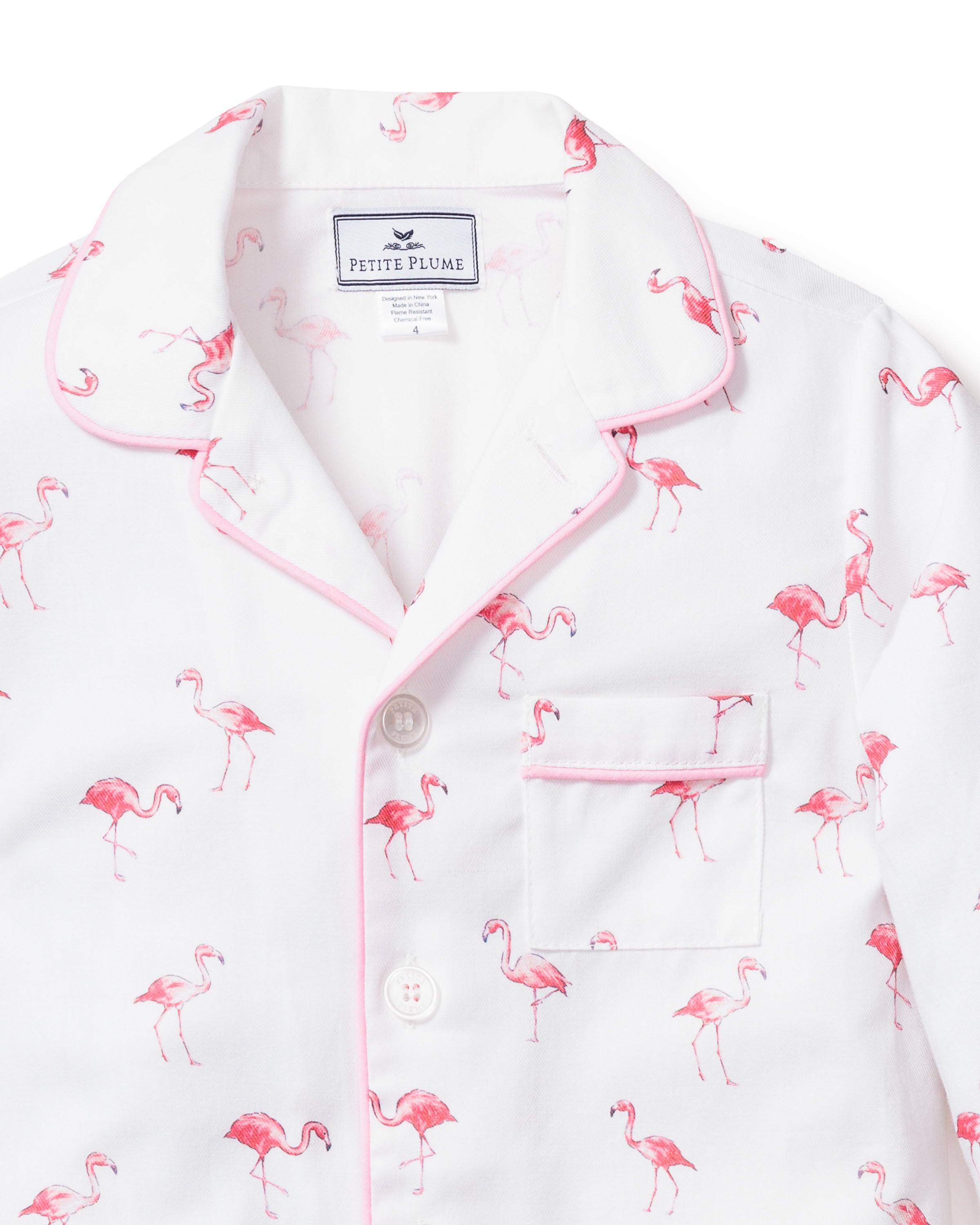 The Kids Twill Pajama Set in Flamingos by Petite Plume features a white top with pink flamingo prints and pink piping, a small chest pocket, and white buttons. Part of a tropical sleepwear set, its crafted from flame retardant cotton for safety.