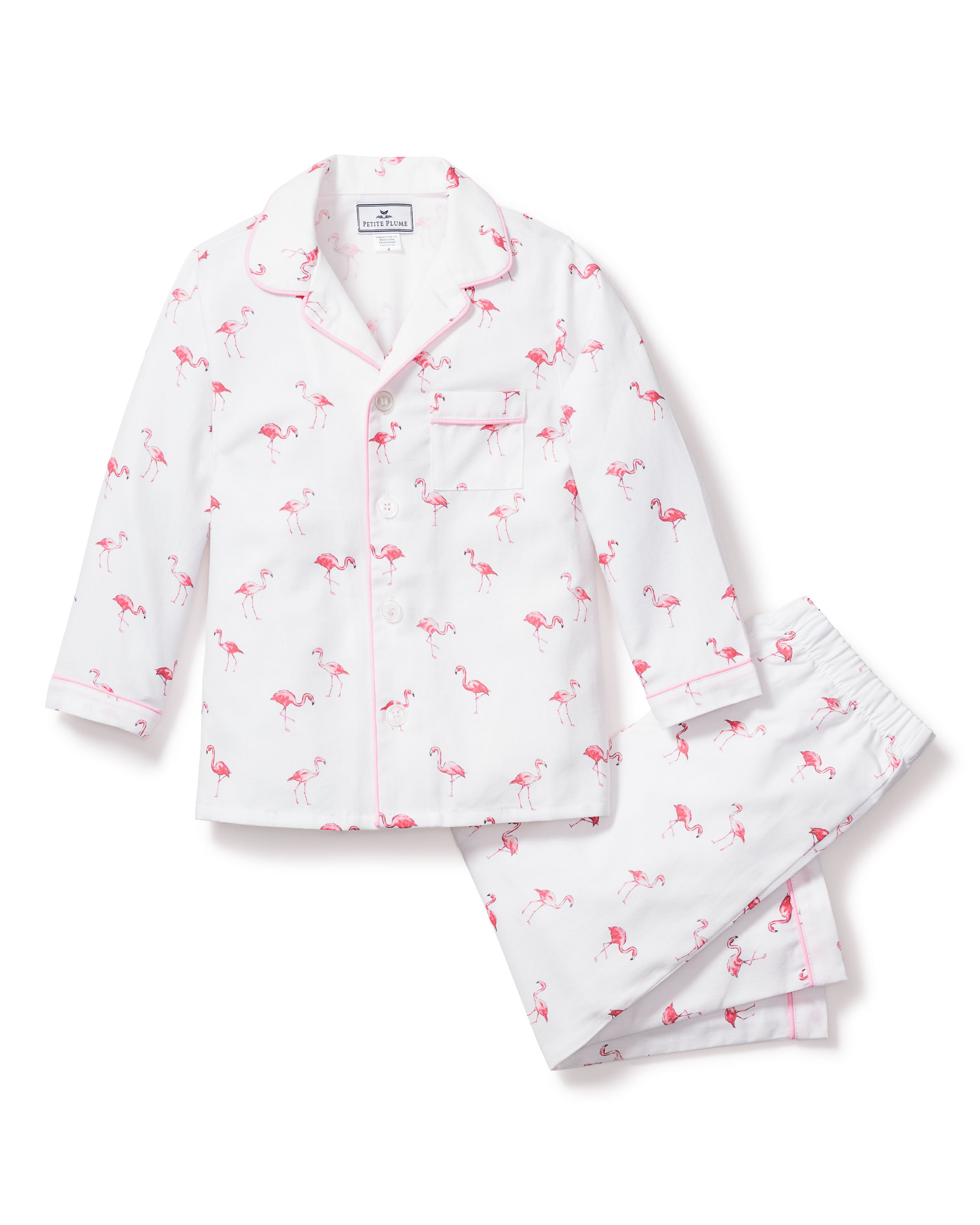 The Kids Twill Pajama Set in Flamingos by Petite Plume is a white tropical sleepwear with a pink flamingo pattern. It includes a long-sleeve button-up top with a chest pocket and matching pants, made from soft, flame retardant cotton for safety and comfort.