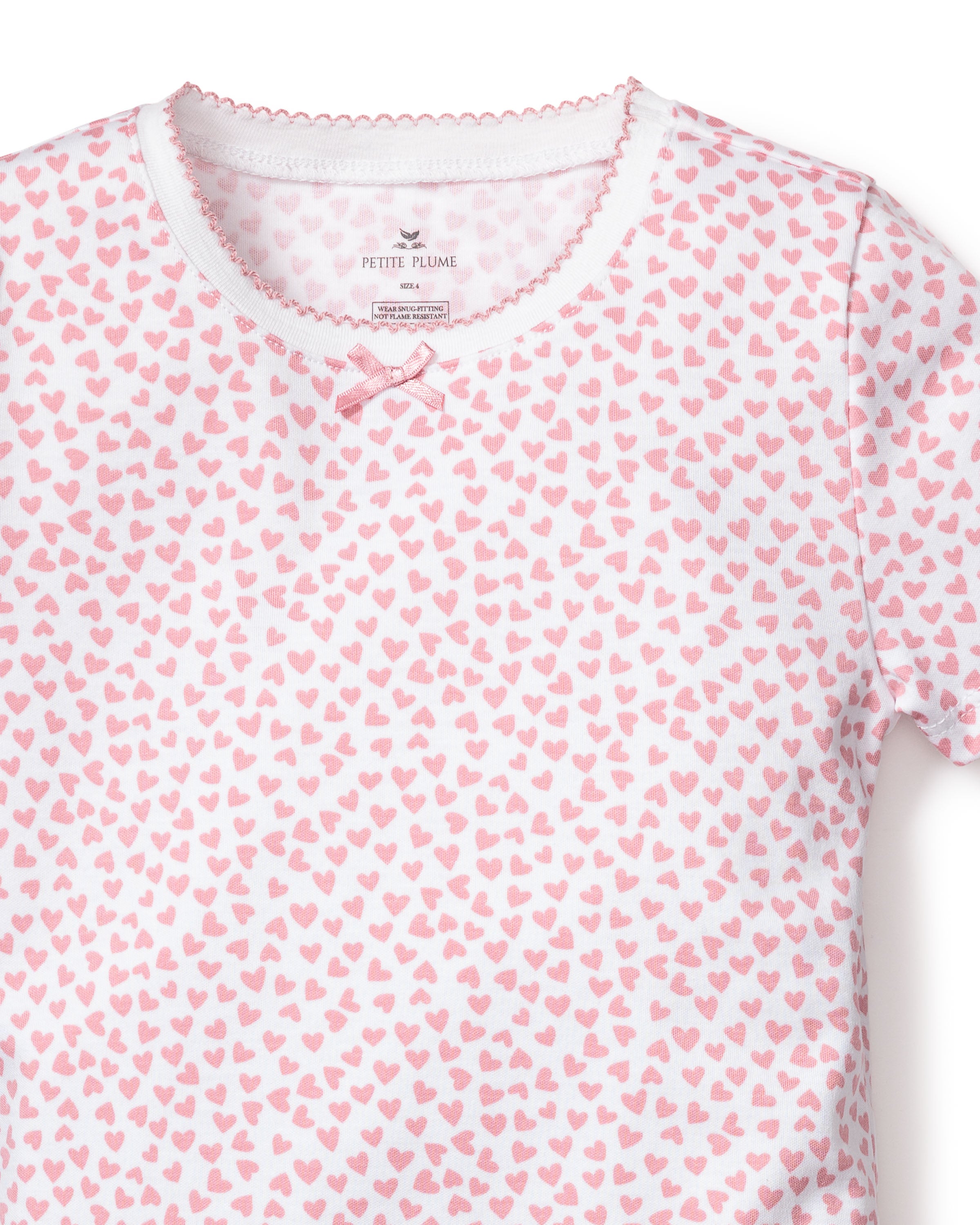 The Kids Pima Snug Fit Pajama Short Set in Sweethearts from Petite Plume features a white T-shirt with a pink heart pattern, a pink bow at the neckline, and scalloped trim. Made from soft Pima Cotton Sleepwear, its ideal for childrens luxury sleepwear. The fabric label inside shows brand details.