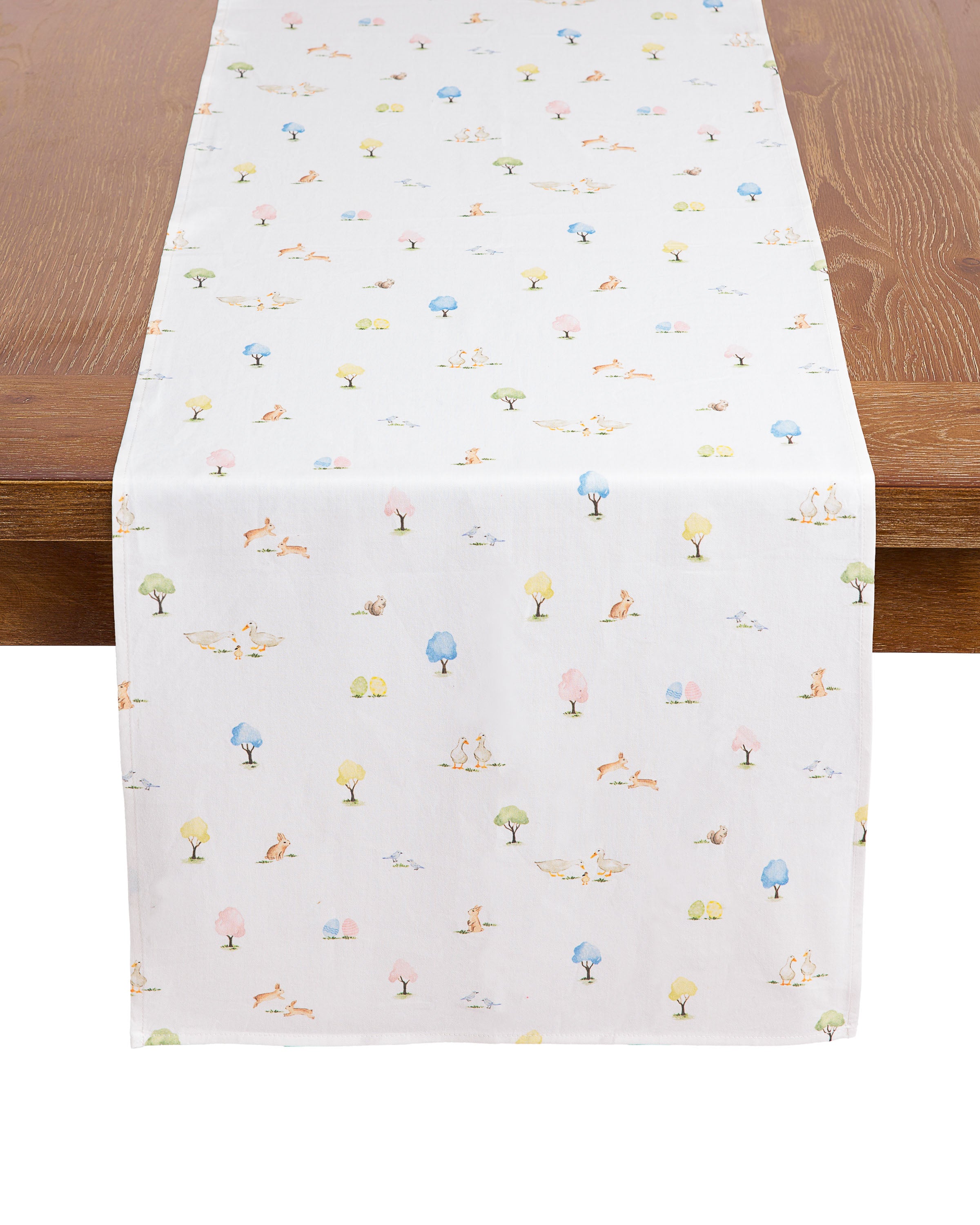 The Easter Gardens Table Linen by Petite Plume features a white table runner made from 100% cotton with vibrant tree and animal illustrations in green, blue, pink, and yellow, including charming birds and foxes, for a whimsical touch to your wooden table.