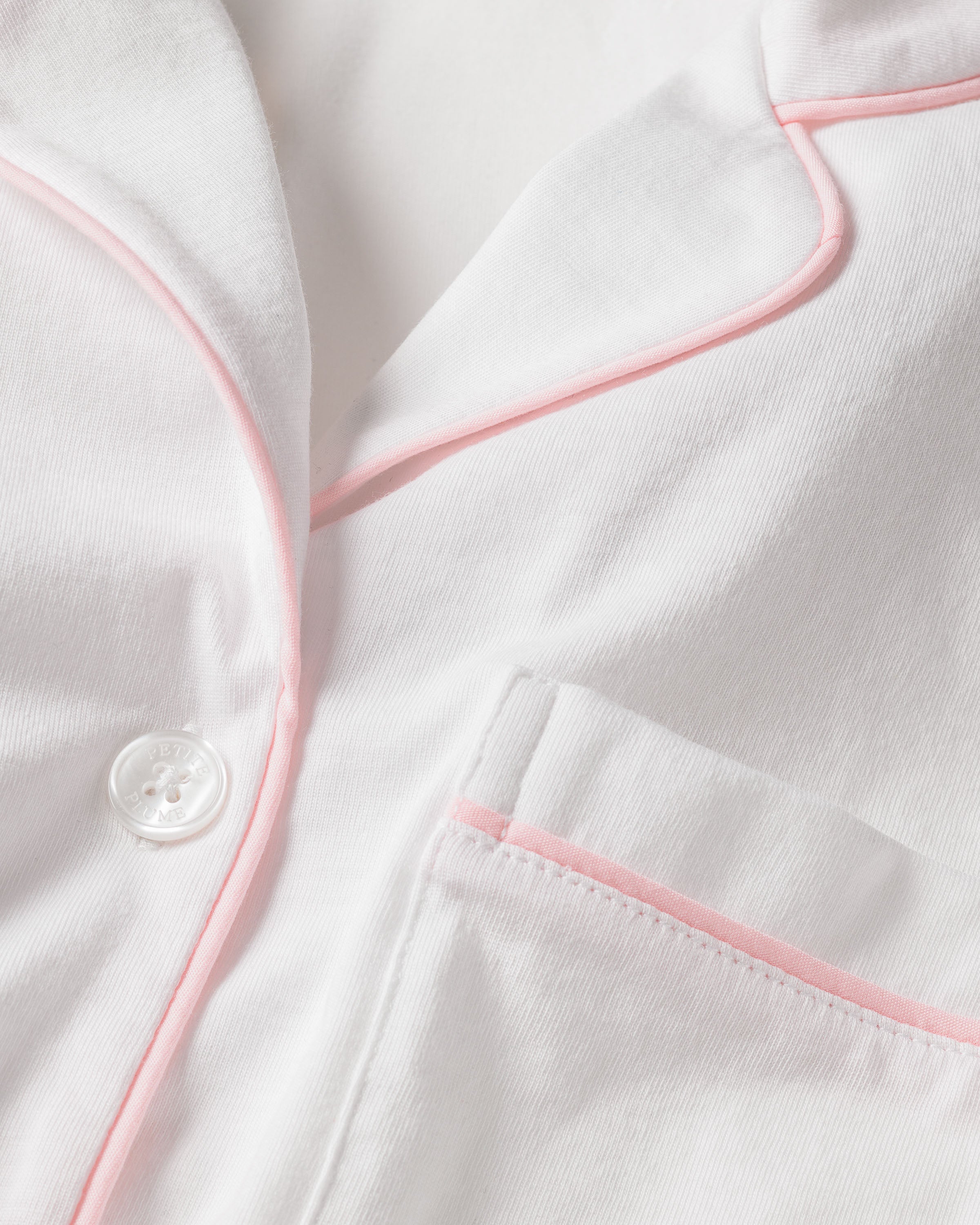 A close-up of the Womens Pima Pajama Short Set in White with Pink Piping by Petite Plume shows a white fabric with pink trim, button, and pocket. The soft texture hints at luxury pajamas crafted from Peruvian Pima cotton.