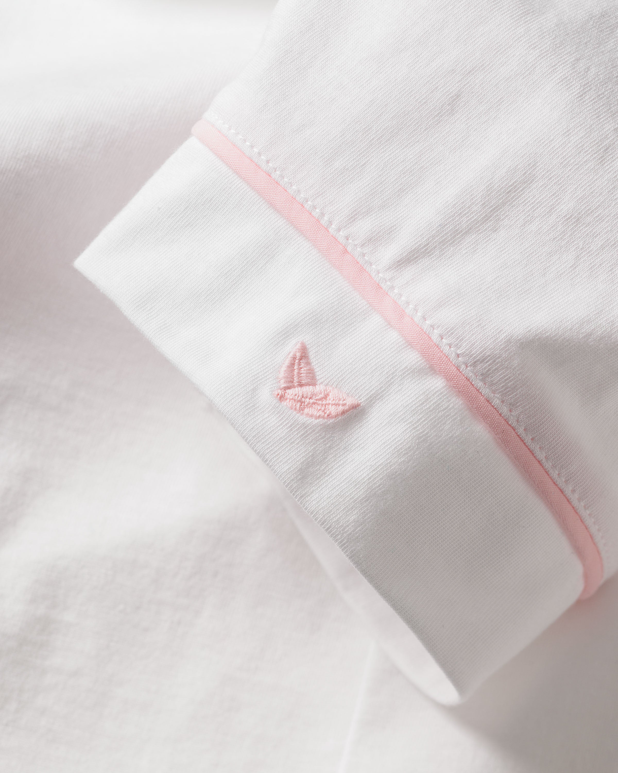 Close-up of Petite Plumes Womens Pima Pajama Short Set in White with Pink Piping, displaying a sleeve crafted from Peruvian Pima cotton. A pink embroidered logo is near the cuff, and a thin pink line accents the edge against the crisp white fabric.