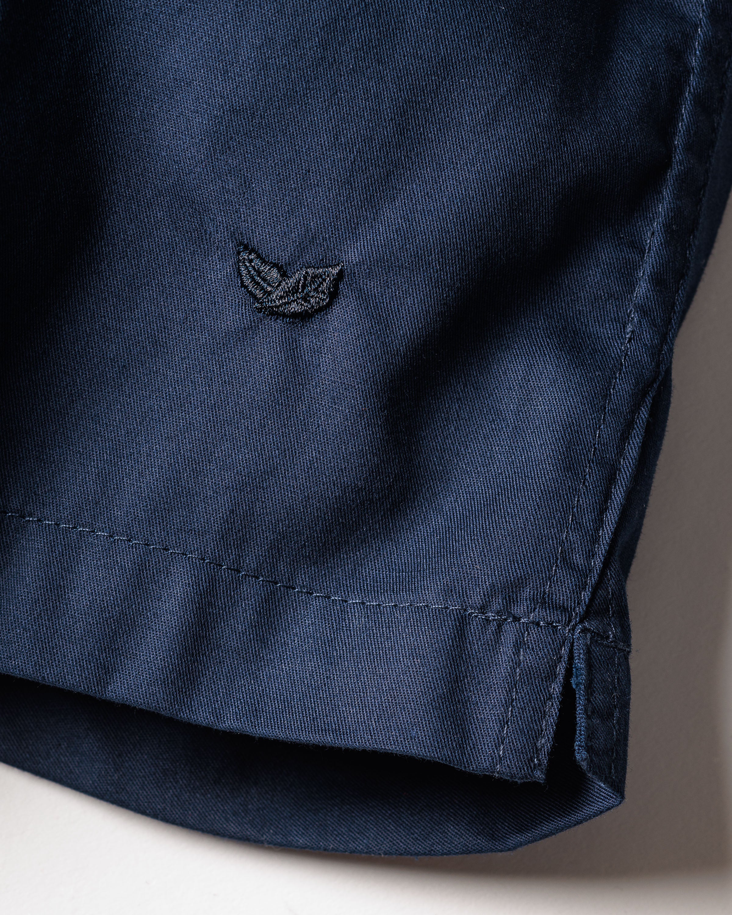 Close-up of a navy fabric from Petite Plumes Mens Twill 3 Pack Boxers, featuring an embroidered logo with two leaves or wings and a neatly finished hem. Made from woven cotton twill, the texture is soft and slightly matte.
