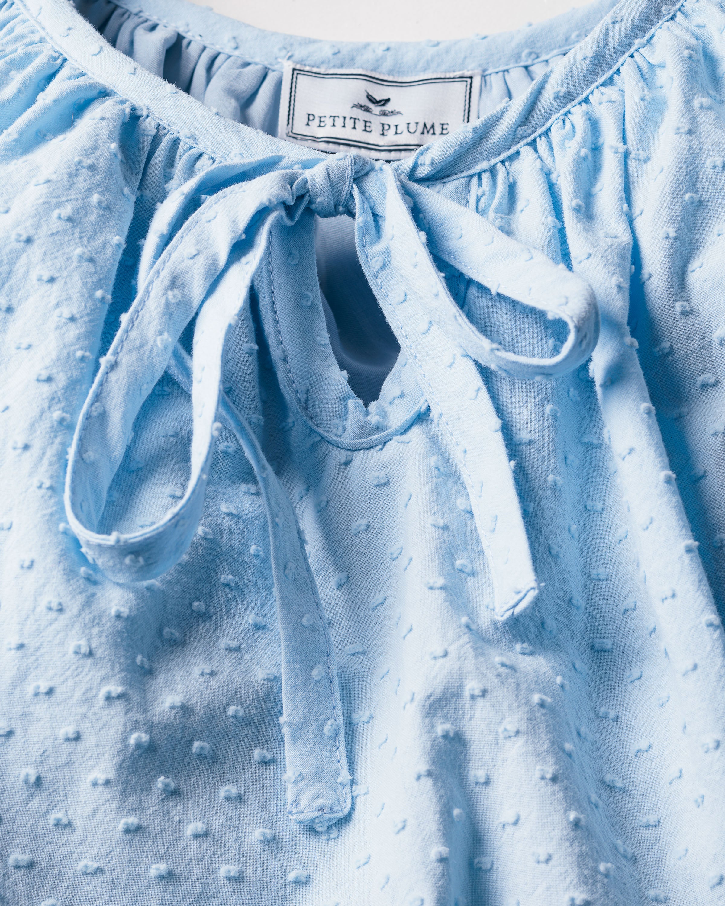 Close-up of a textured light blue Womens Swiss Dots Celeste Short Set by Petite Plume, adorned with Swiss dots and featuring an elegant keyhole neckline with a bow at the top.
