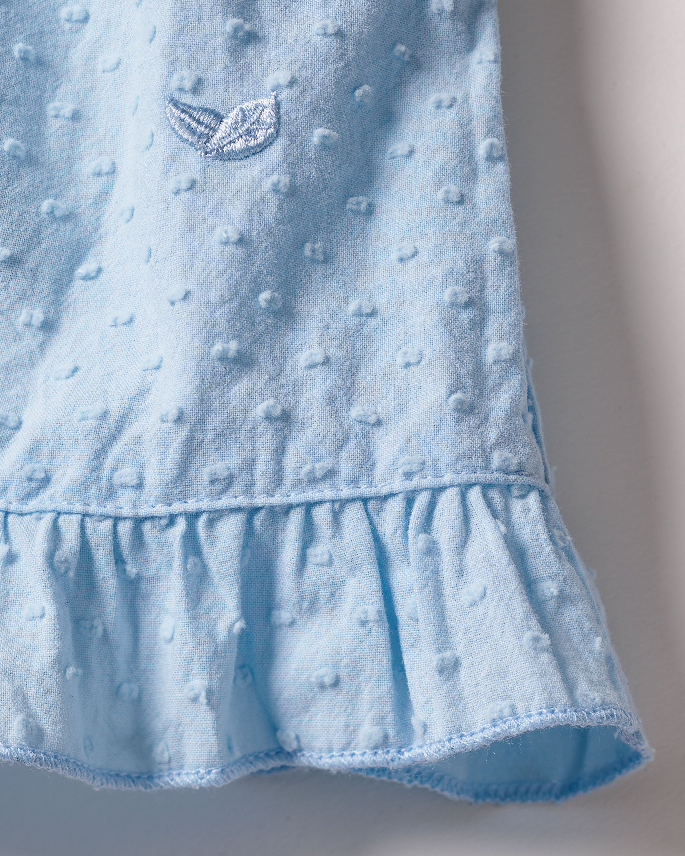 The Womens Swiss Dots Celeste Short Set in Blue by Petite Plume showcases a light blue cotton Swiss Dot fabric with an embossed dot pattern, small embroidered leaf design, and a ruffled edge at the bottom, adding texture and detail for luxurious sleepwear.