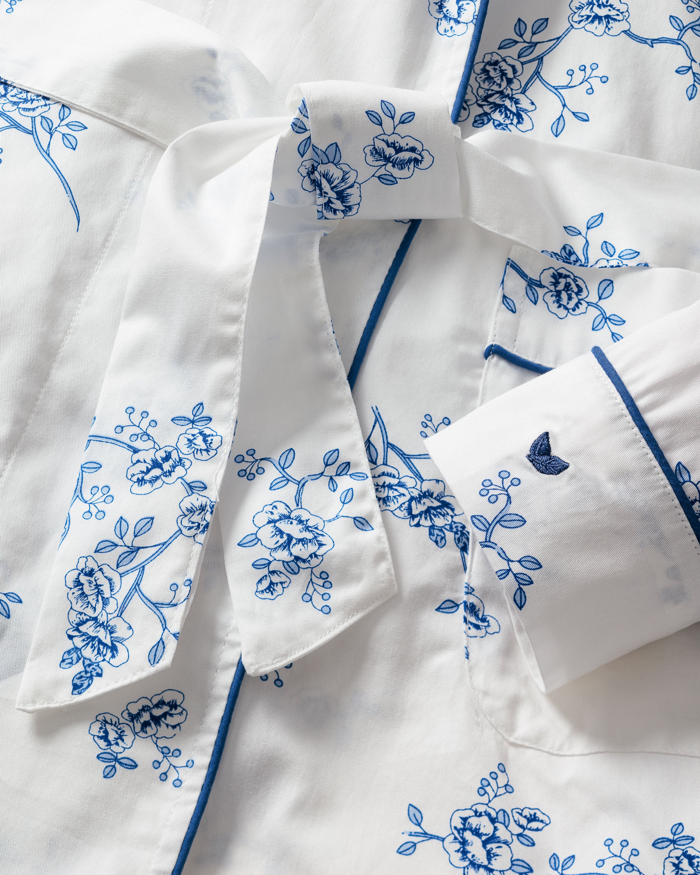Close-up of a white fabric with blue floral prints: Kids Twill Robe in Indigo Floral by Petite Plume. Made from premium cotton, it features a neatly folded collar and cuffs with dark piping, exuding classic elegance with intricate flower patterns.
