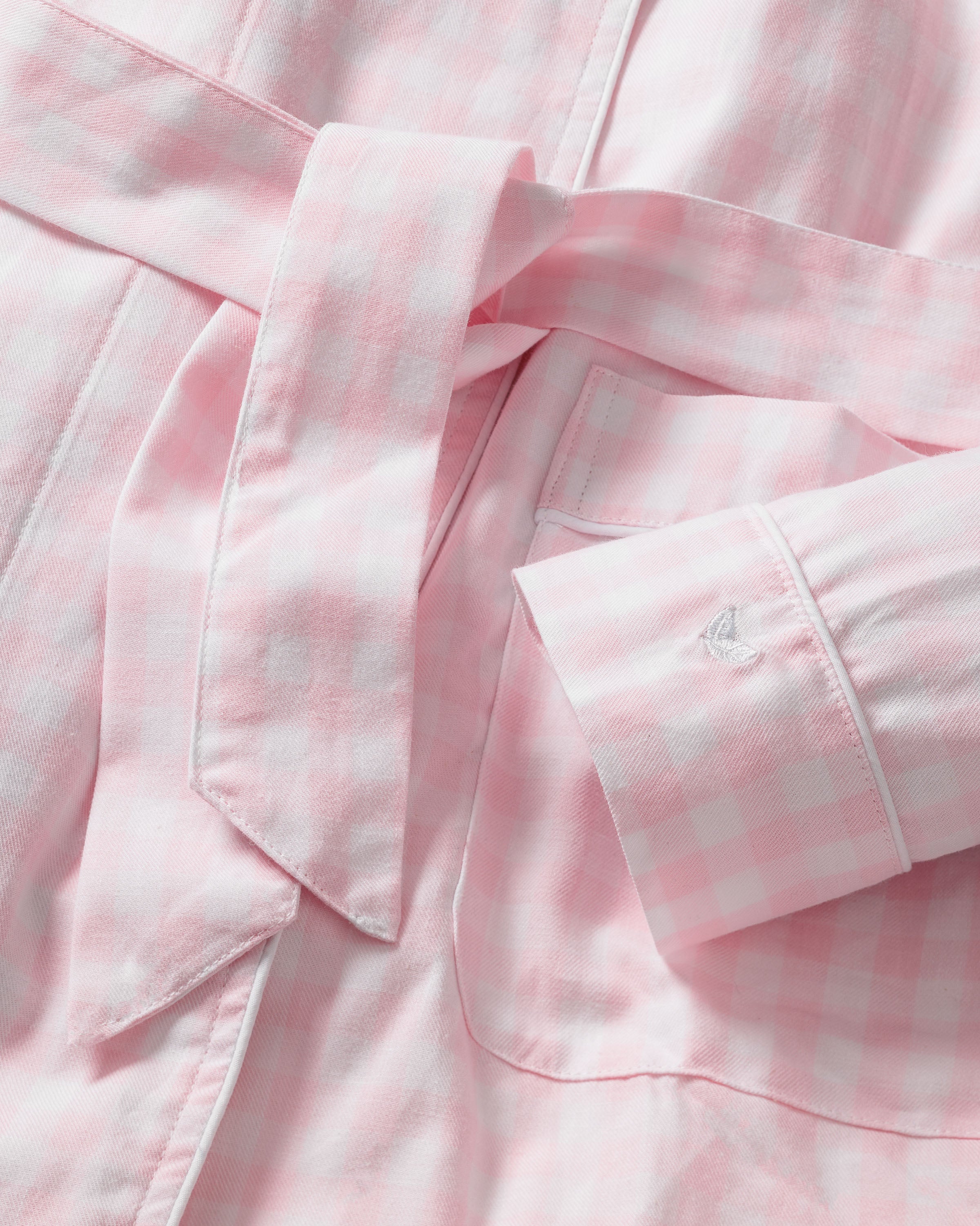 A close-up of Petite Plumes Kids Twill Robe in Pink Gingham shows its soft, textured fabric, reminiscent of classic childrens sleepwear. The robe features a folded cuff with stitched letters and a tied belt across the light pink and white gingham pattern.
