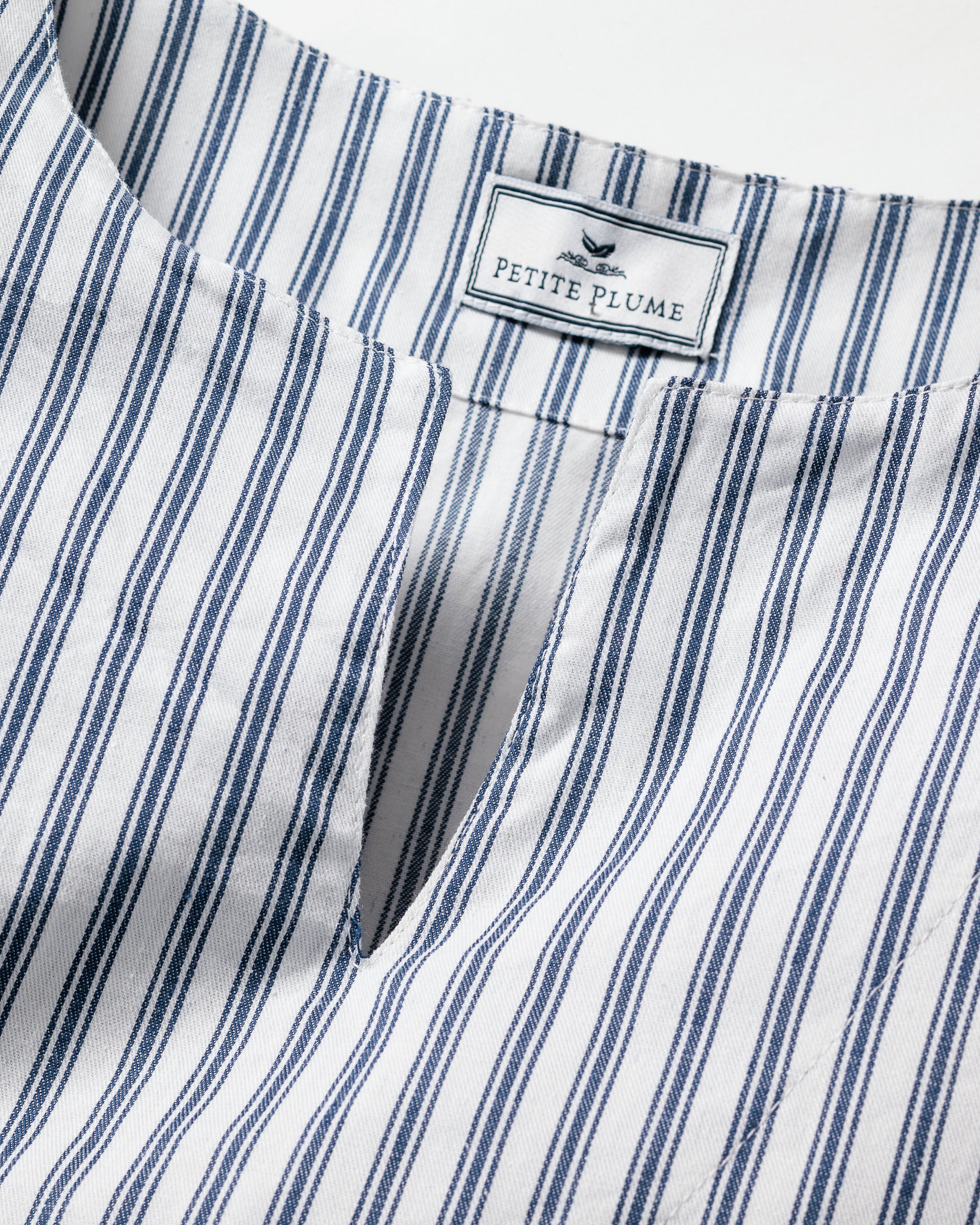 Close-up of a breathable cotton fabric with vertical navy blue and white stripes, featuring a notch neckline. A label at the back reads Petite Plume with a bird logo. Ideal for luxurious sleepwear or stylish caftans, this is the Girls Twill Caftan in Navy French Ticking by Petite Plume.