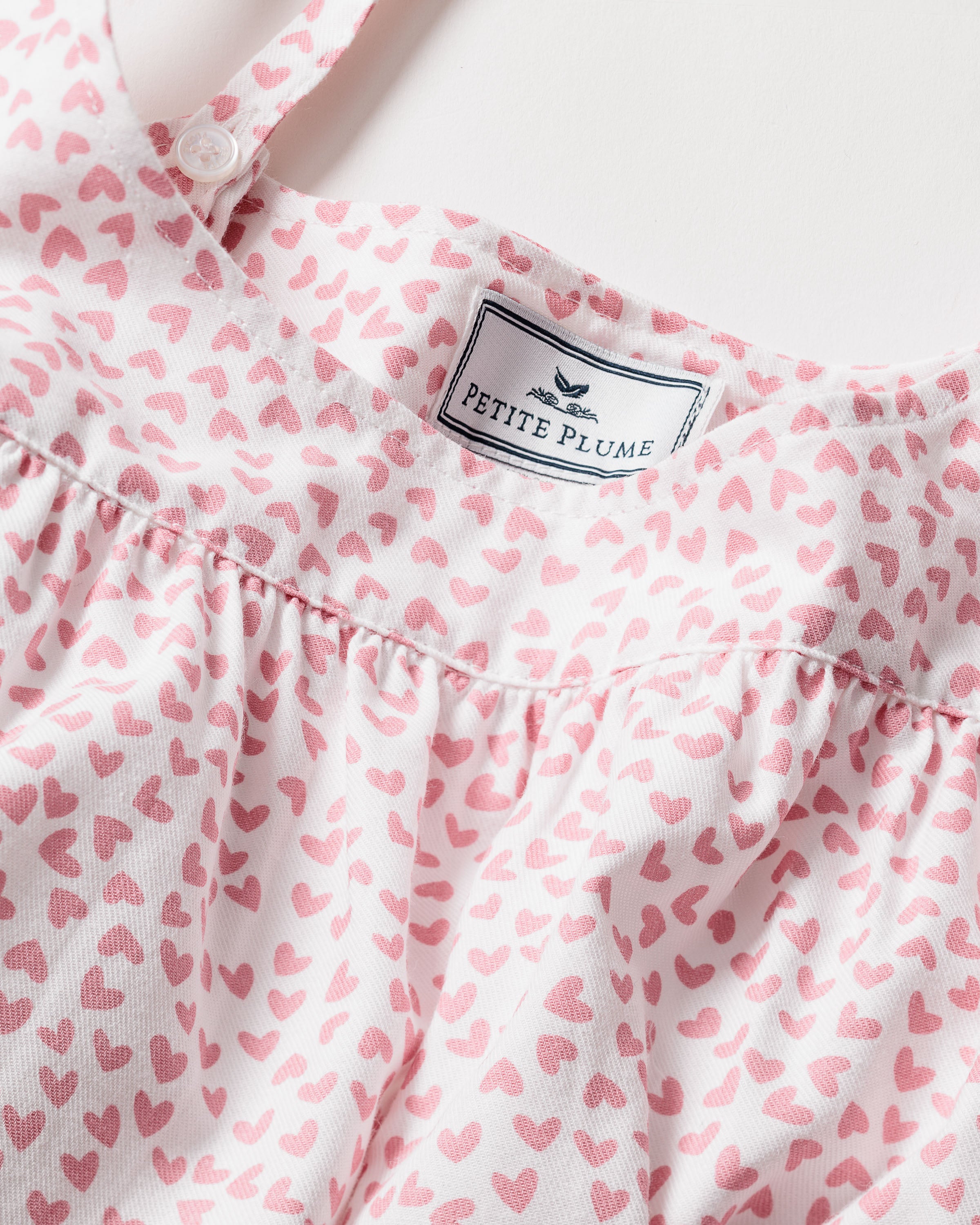Close-up of a garment label displaying the Petite Plume brand with a bird logo, showcasing the Womens Twill Chloé Nightgown in Sweethearts. Made from premium cotton, it features pink heart patterns on white fabric and has a gathered design beneath the label for luxury sleepwear elegance.
