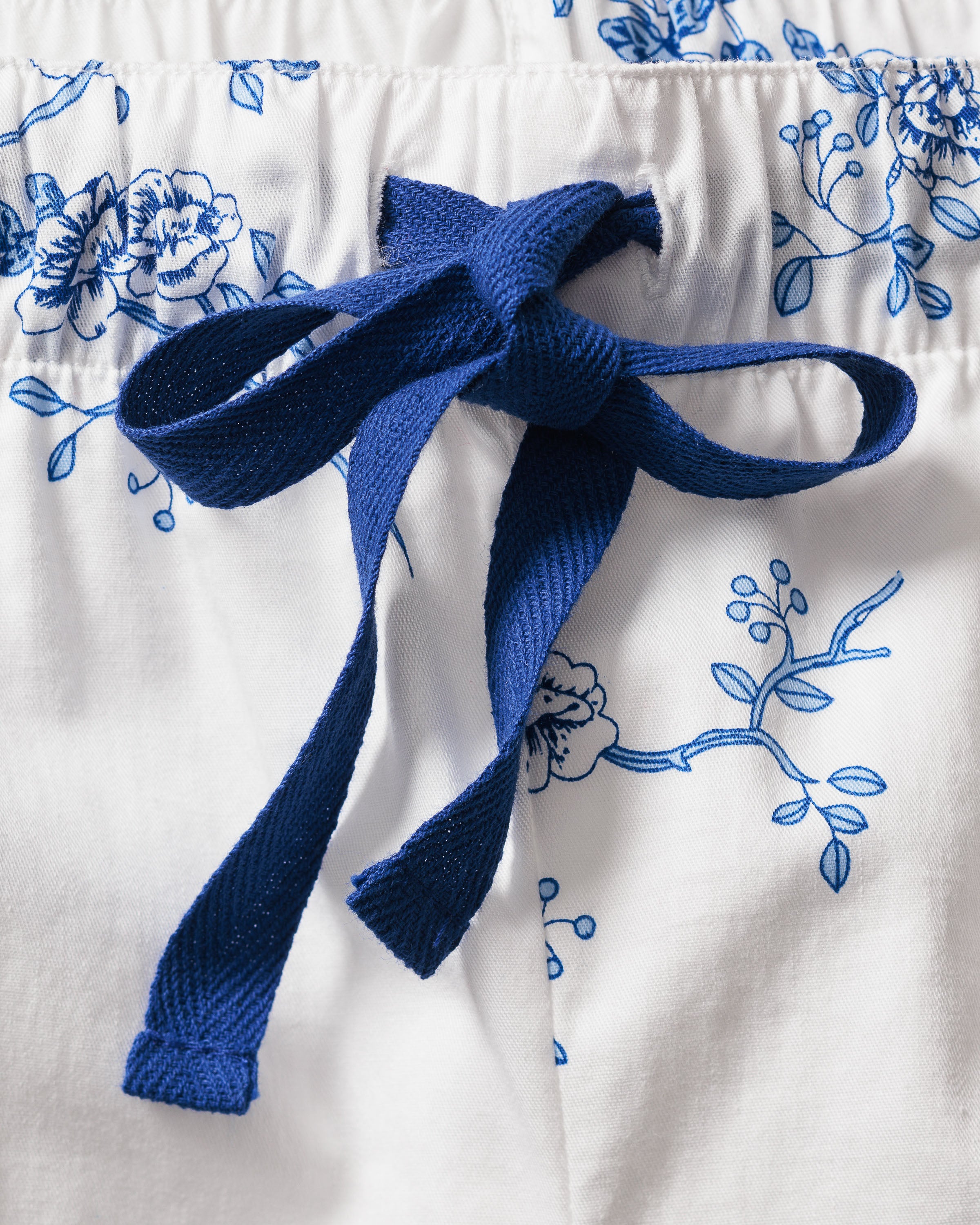 Close-up of the Womens Twill Long Sleeve Short Set in Indigo Floral by Petite Plume, featuring a blue and white floral pattern on premium cotton. The central focus is a navy blue drawstring bow, set against intricate vintage-style blue flowers and branches on this luxurious sleepwear.