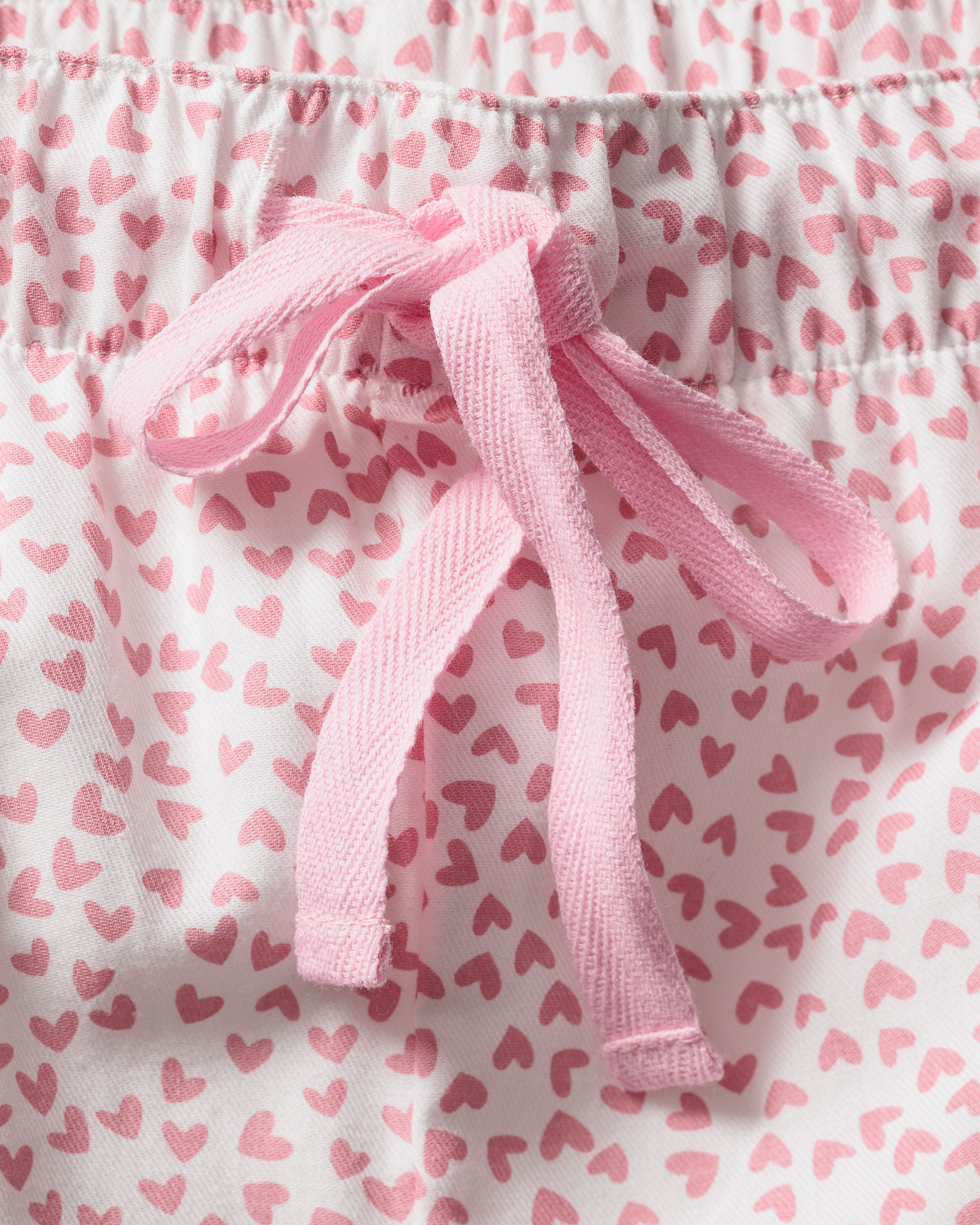 A close-up of the Womens Twill Pajama Set in Sweethearts by Petite Plume highlights a white fabric with small pink hearts and a pink drawstring bow, epitomizing luxury sleepwear. The gathered material suggests pajama shorts or pants made for comfort.