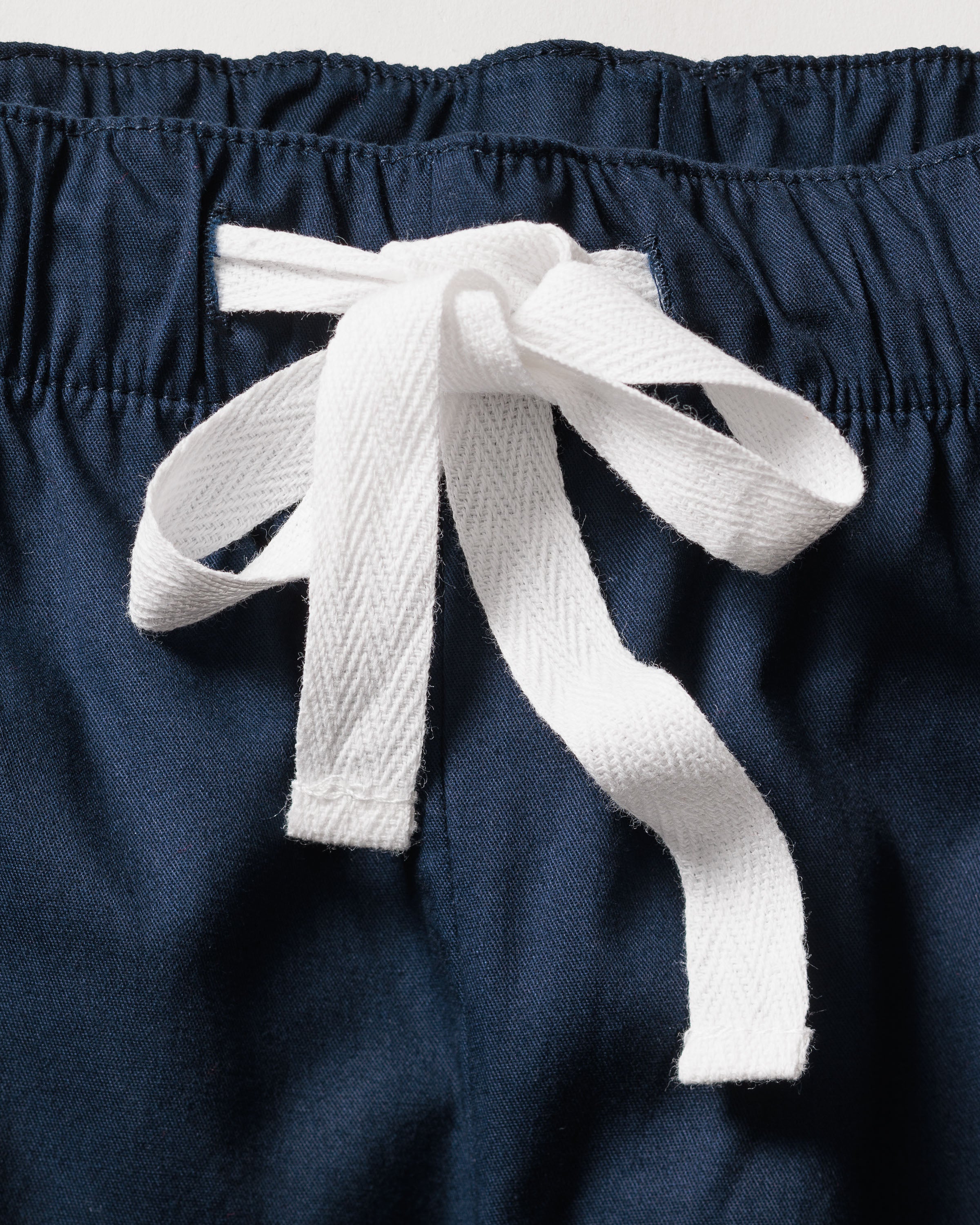 Close-up of Petite Plumes Mens Navy Twill Pajamas, featuring luxurious yarn-dyed cotton with white piping and a white drawstring bow. The fabric is gently gathered at the waistband, accentuating the color contrast.