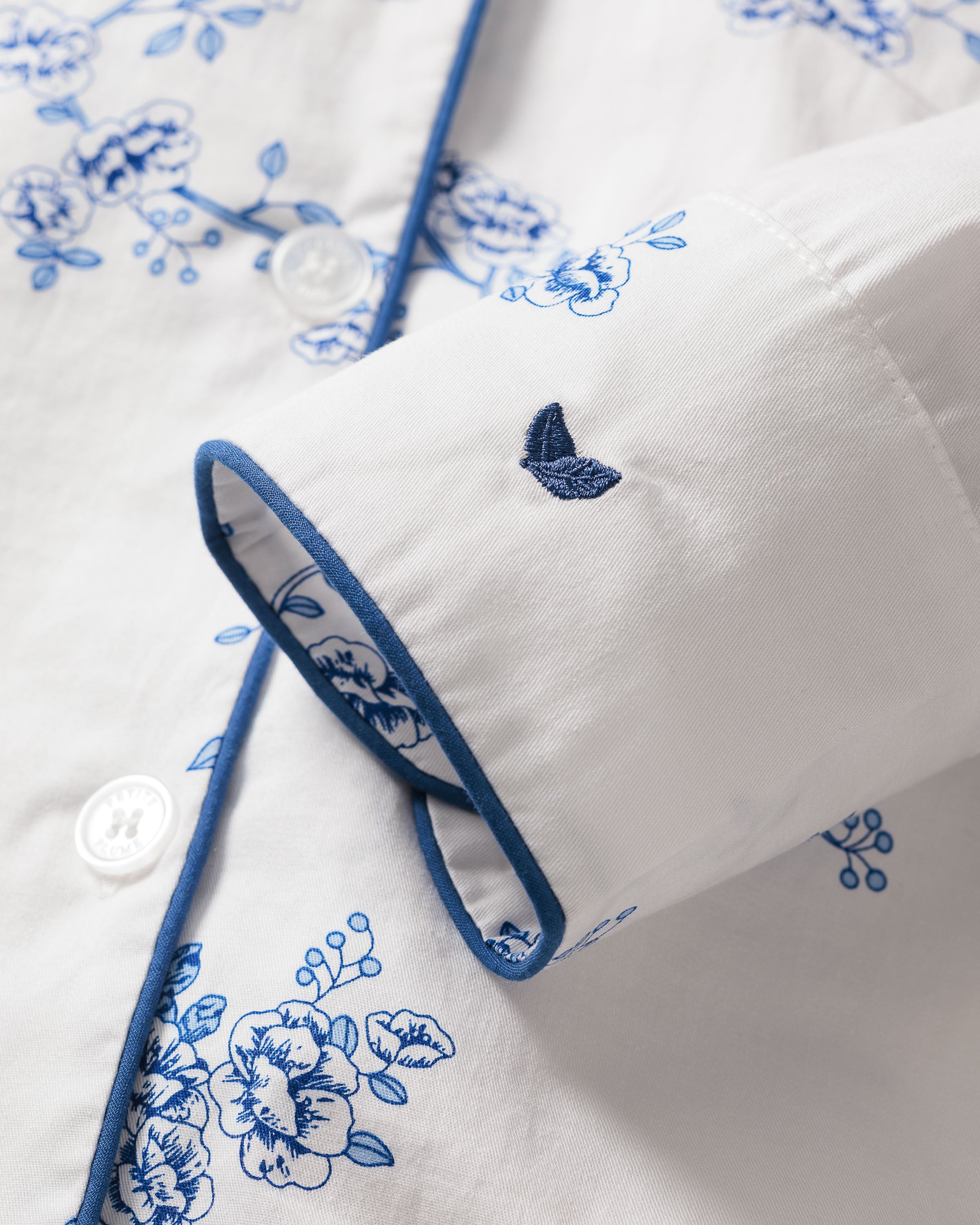 Close-up of the Womens Twill Long Sleeve Short Set in Indigo Floral by Petite Plume, made from top-quality cotton with blue floral patterns, blue piping on the button placket and sleeve edges, an embroidered butterfly logo on the cuff, and white buttons completing the vintage design.