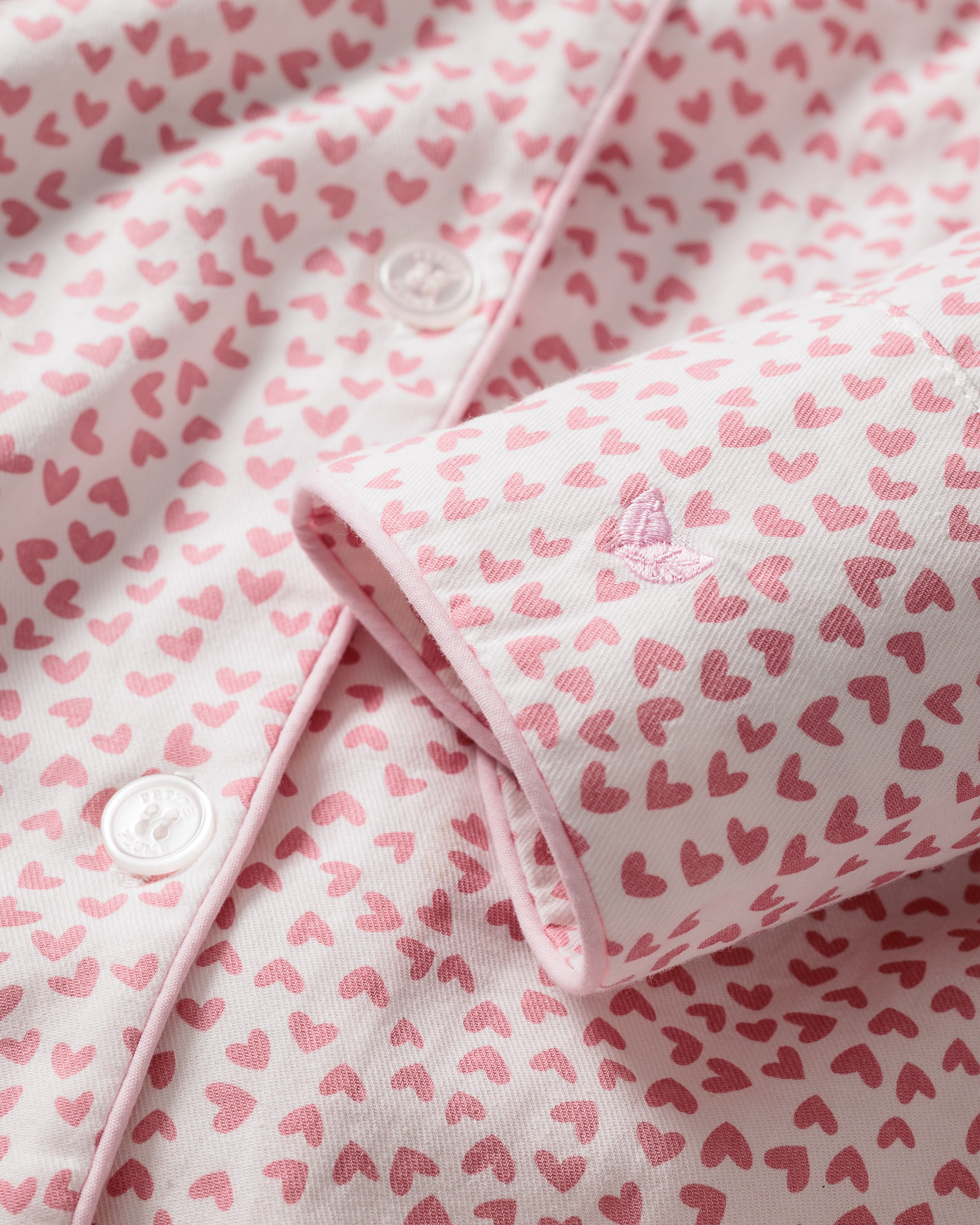 Close-up of Petite Plumes Womens Twill Pajama Set in Sweethearts, featuring pink hearts and embroidery on a light background. The button closure, visible stitching, and partially rolled sleeve with extra heart embroidery enhance this luxury sleepwears charm.