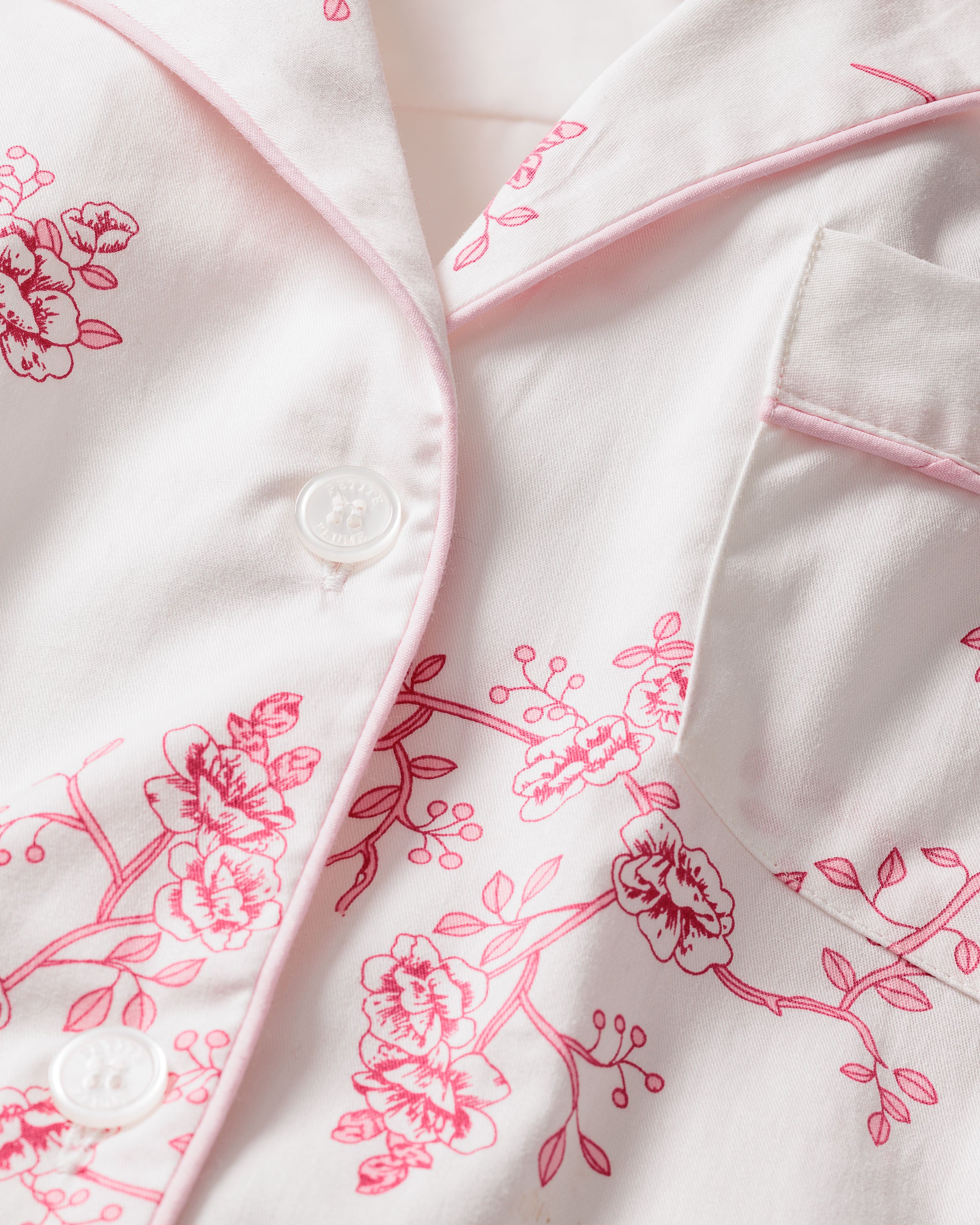 Close-up of the Kids Twill Pajama Set in English Rose Floral by Petite Plume. This classic set features a luxurious white top with pink floral patterns, piping, button-up front with visible buttons, and a small chest pocket. The soft fabric ensures a comfortable nights sleep.